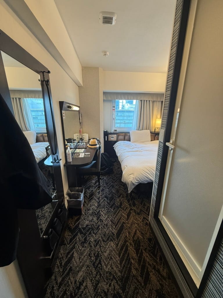 Compact hotel room at APA Hotel Osaka Umeda Eki Tower, featuring a double bed, desk, mirror, luggage and a window overlooking central Osaka.
