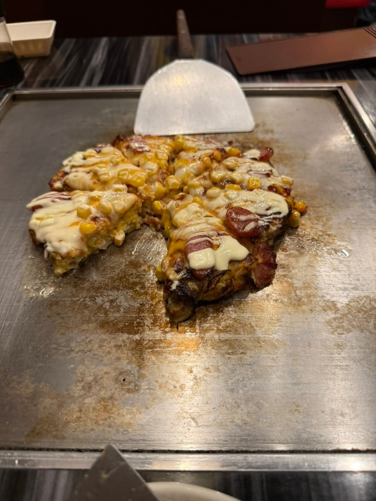 Okonomiyaki cooking on a teppan grill in Osaka, topped with sweetcorn, sliced sausage and melted cheese, with a metal spatula resting behind the dish.