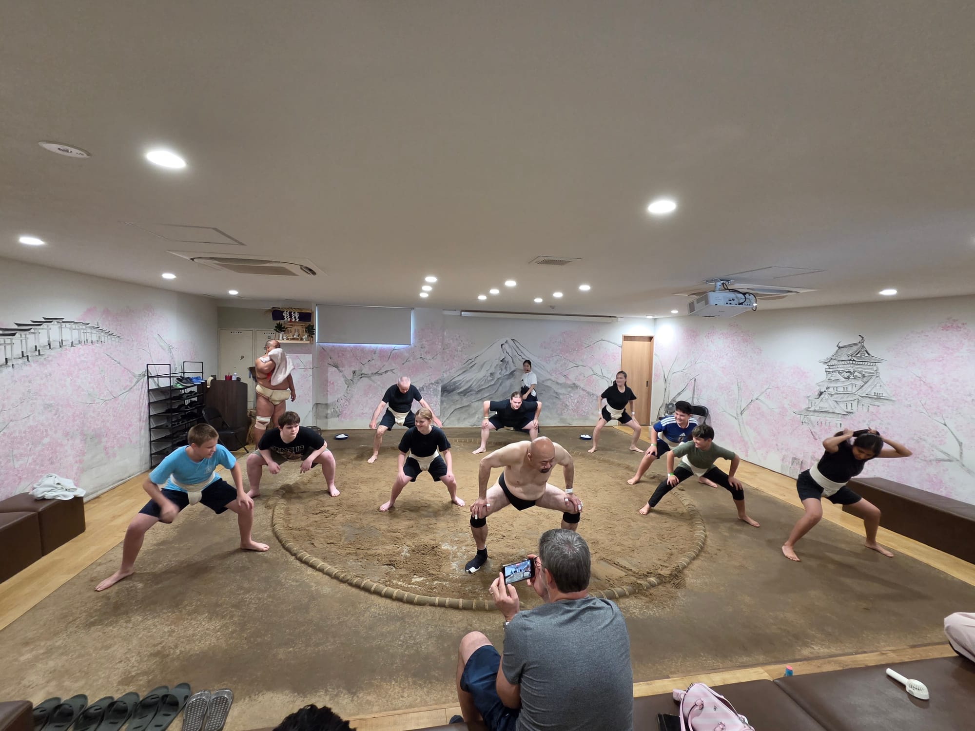 Participants practise sumo wrestling techniques in a training ring, following an instructor's movements inside a dojo decorated with murals of Mount Fuji and Osaka Castle.
