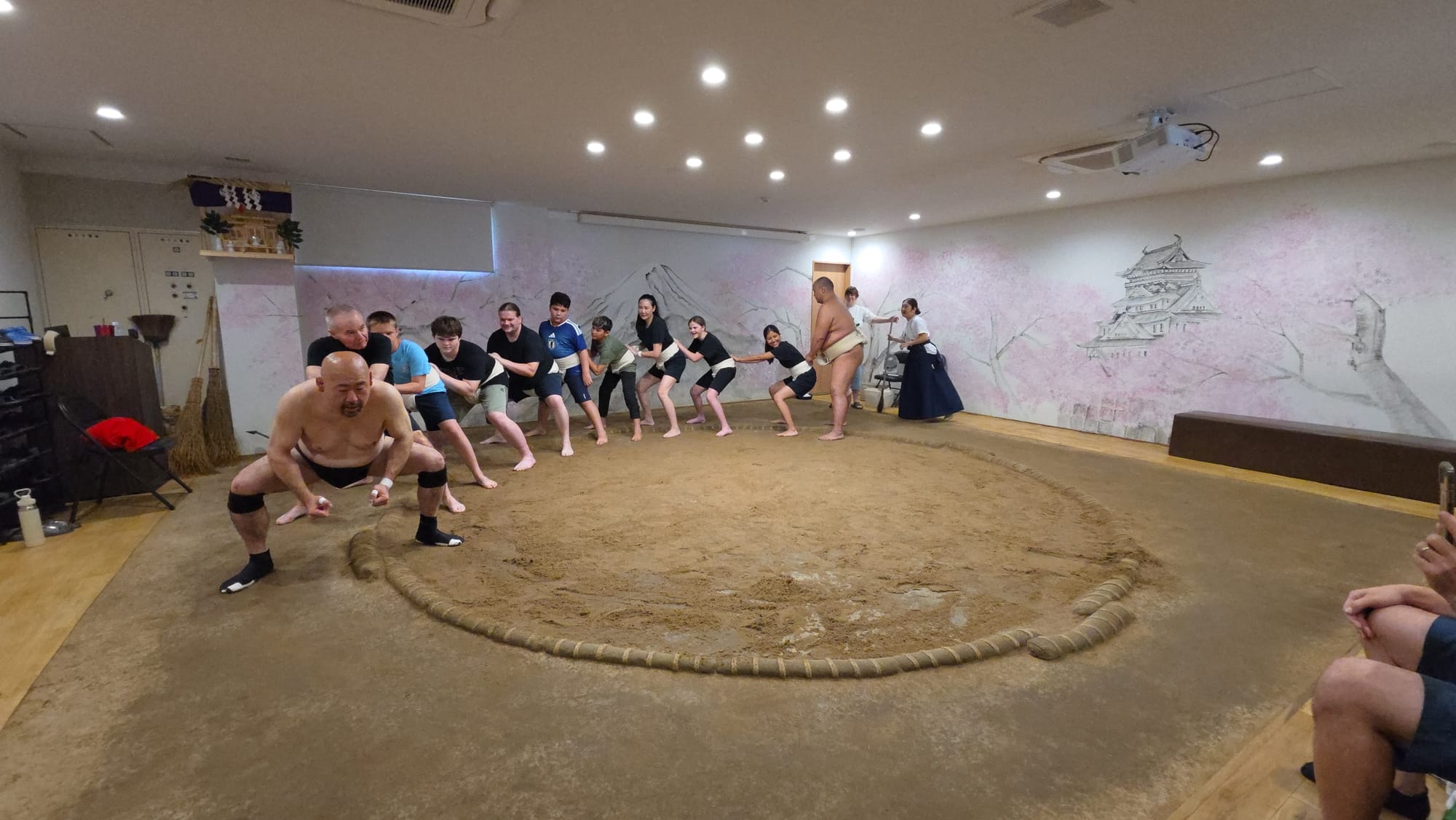 Participants practise a sumo pushing exercise inside a dojo in Osaka, following instructions from a former wrestler around the clay ring during a hands-on training session.