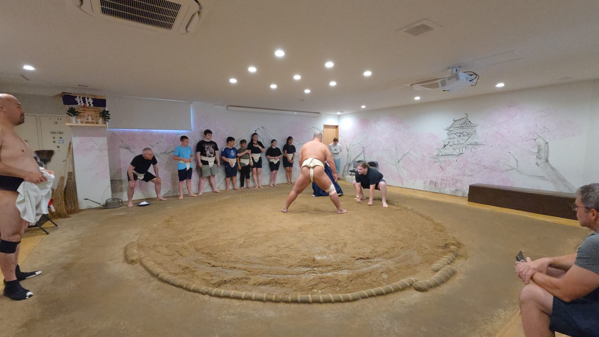 Participant prepares for a sumo pushing exercise inside a dojo in Osaka, facing a former wrestler in the ring while other visitors watch and await their turn.