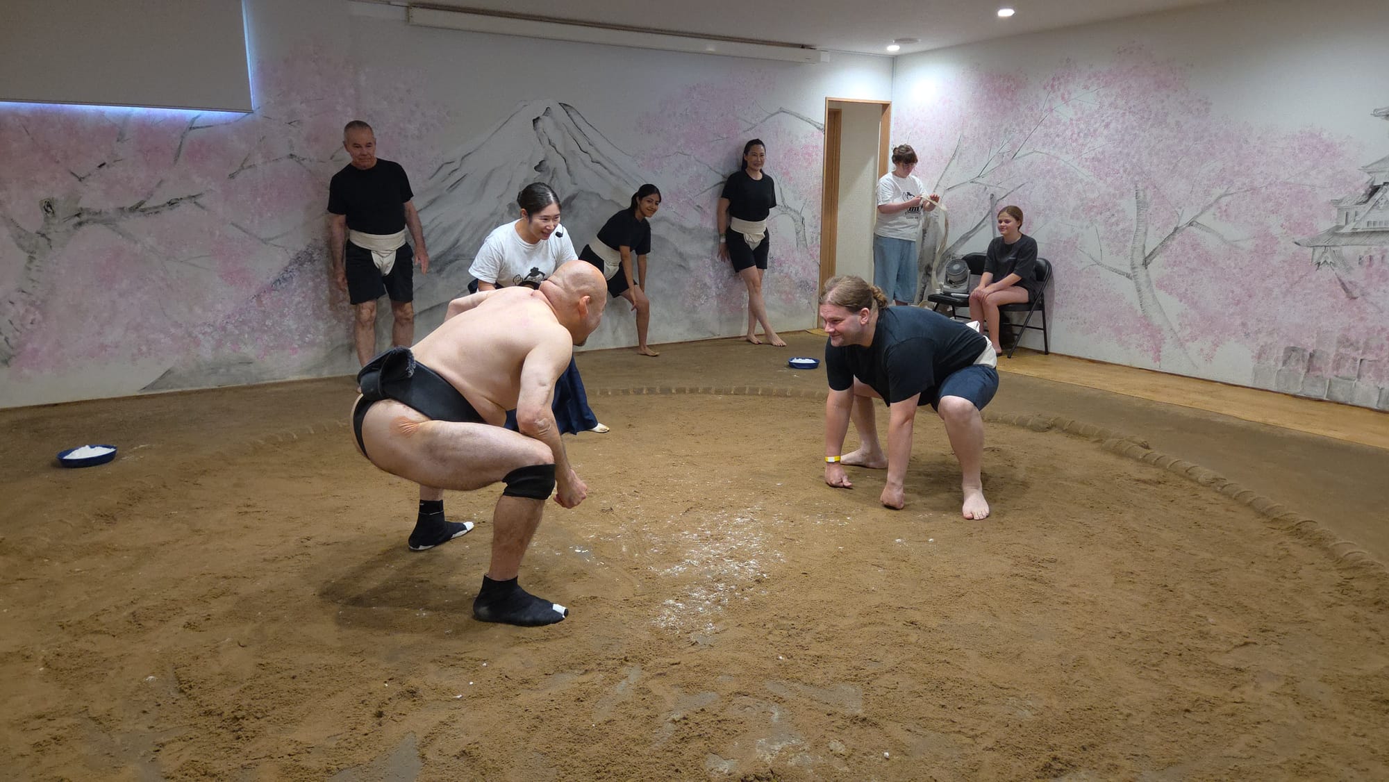 Participant and former sumo wrestler crouch facing each other in a sumo ring in Osaka, preparing for a practice bout while other visitors watch from around the dojo.