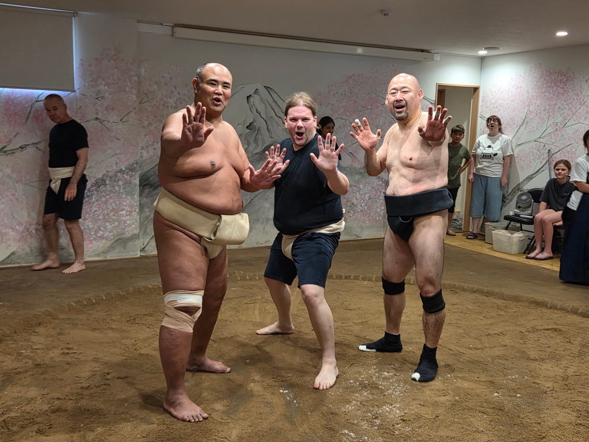 Participant poses with two former sumo wrestlers inside a dojo in Osaka after a training session, standing together in the ring with hands raised towards the camera.