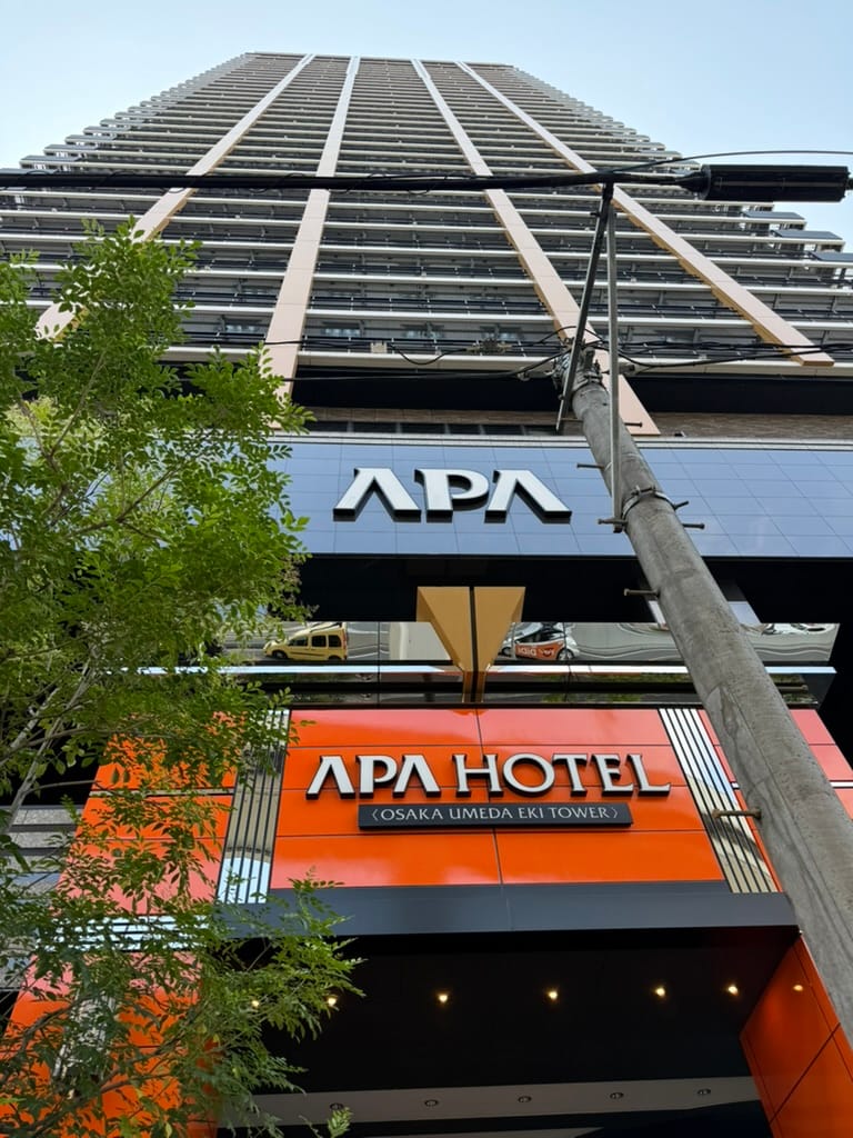 APA Hotel Osaka Umeda Eki Tower rises above the street in Osaka, viewed from below, with the hotel's distinctive orange entrance sign dominating the foreground.