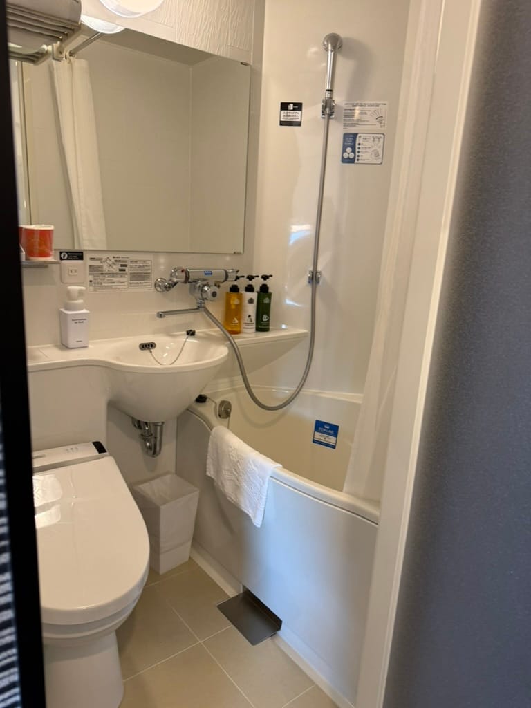 Compact bathroom at APA Hotel Osaka Umeda Eki Tower, featuring a Japanese toilet, small washbasin, shower and deep bathtub arranged within a space-saving layout.