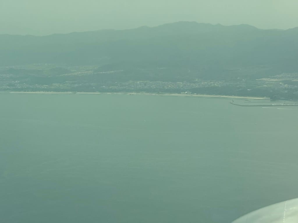 View from an aircraft approaching Japan, looking across Osaka Bay towards Misaki, with a long coastline, harbour facilities and mountains visible through light haze.
