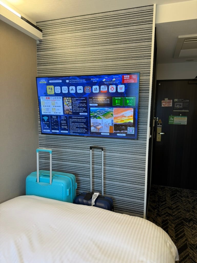 Hotel room at APA Hotel Osaka Umeda Eki Tower, featuring a wall-mounted smart television, luggage cases beside the bed and information displayed on the screen.