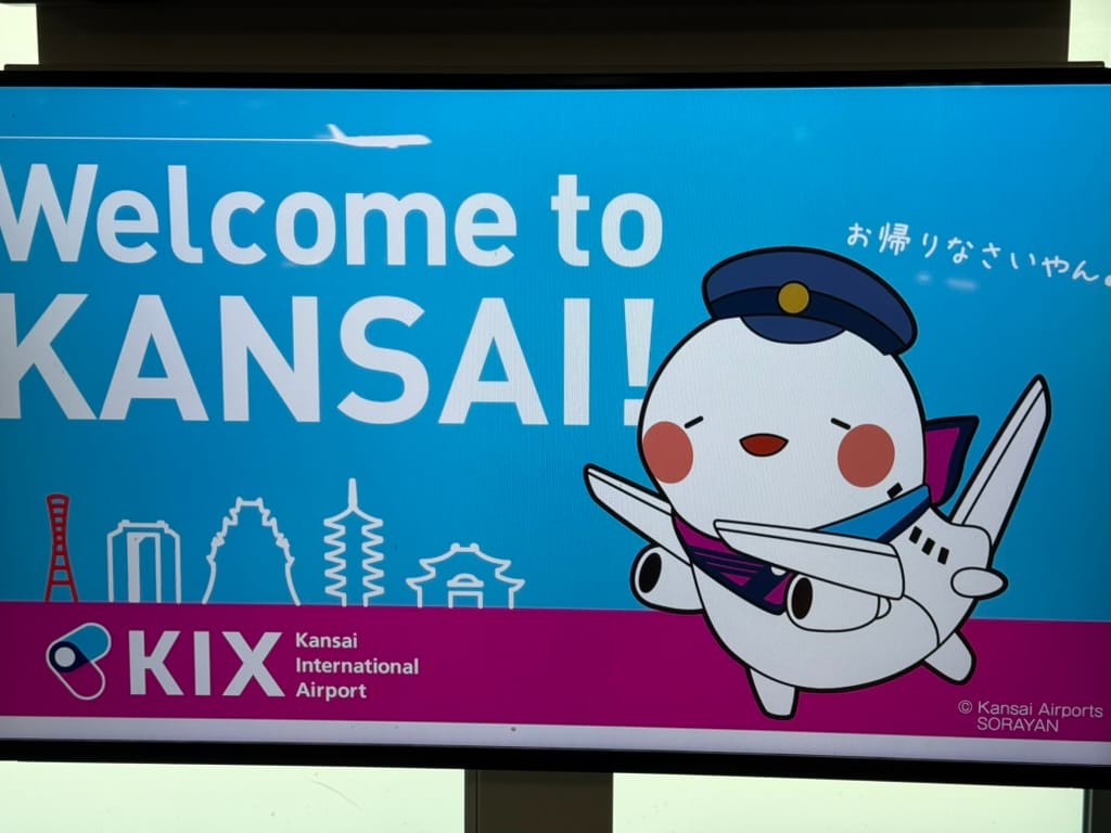 Welcome sign at Kansai International Airport featuring the airport mascot Sorayan dressed as an aeroplane, with landmarks of the Kansai region illustrated in the background.