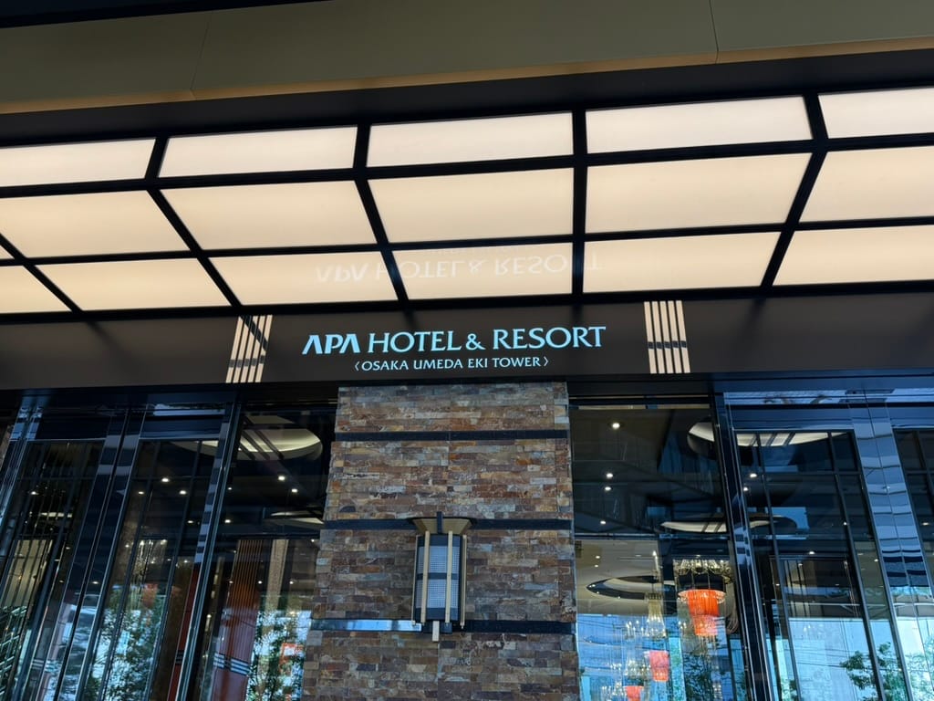 Entrance to APA Hotel & Resort Osaka Umeda Eki Tower, featuring illuminated signage, glass doors, stone-clad columns and a modern canopy above the main entrance.