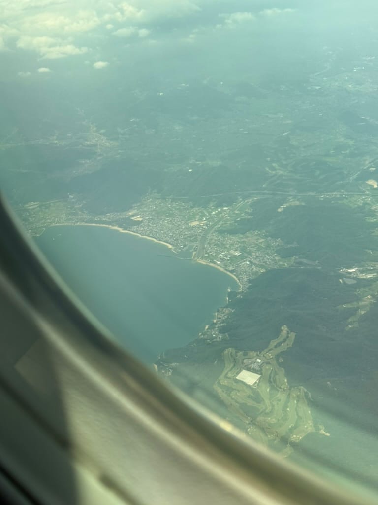View from an aircraft window approaching Japan, showing the coastline of Shikoku with bays, hills and settlements visible below through light haze.