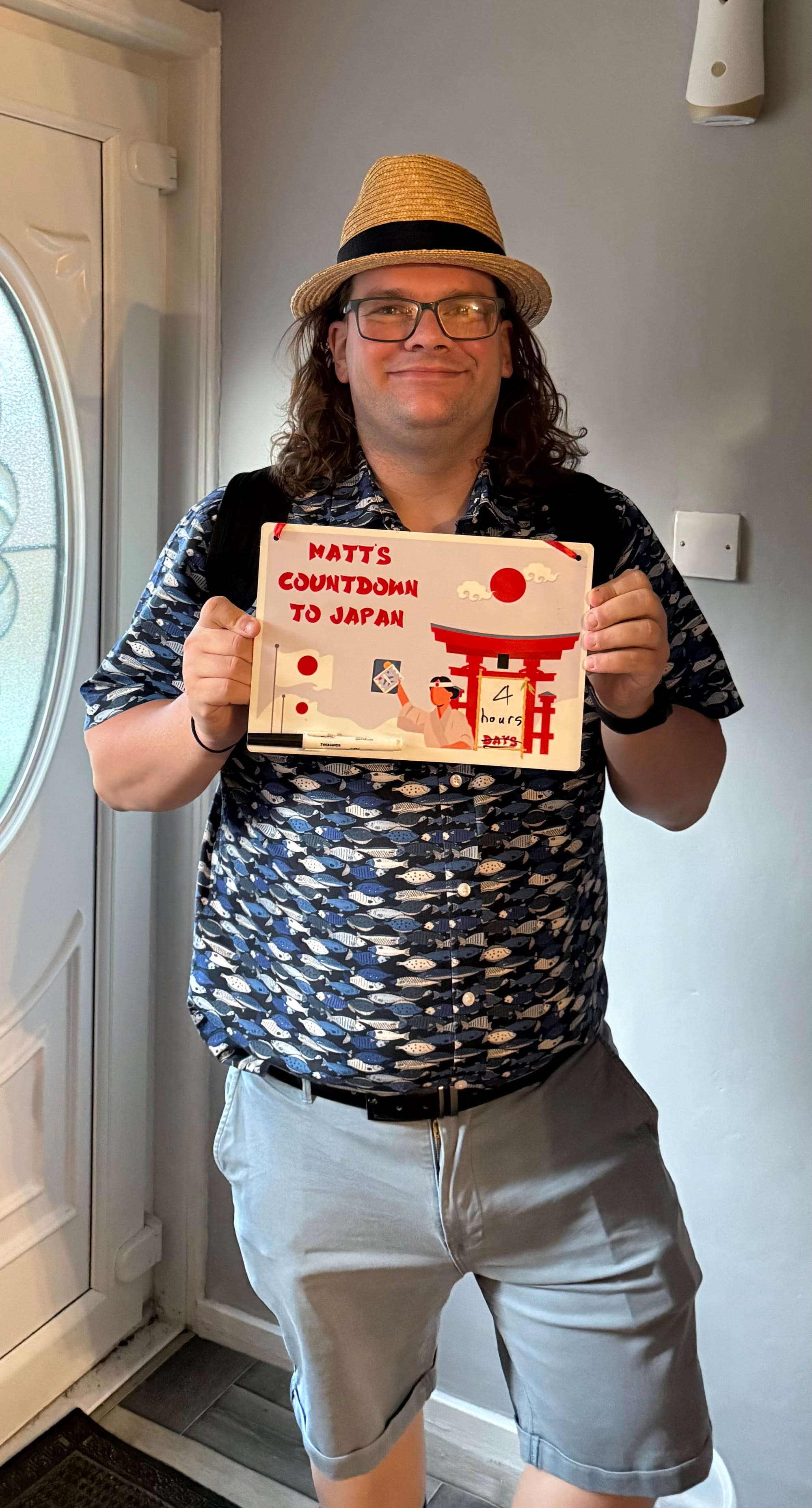 Traveller wearing a straw hat and patterned shirt holds a "Matt's Countdown to Japan" sign beside a front door, with Japanese-themed artwork and a four-hour countdown.