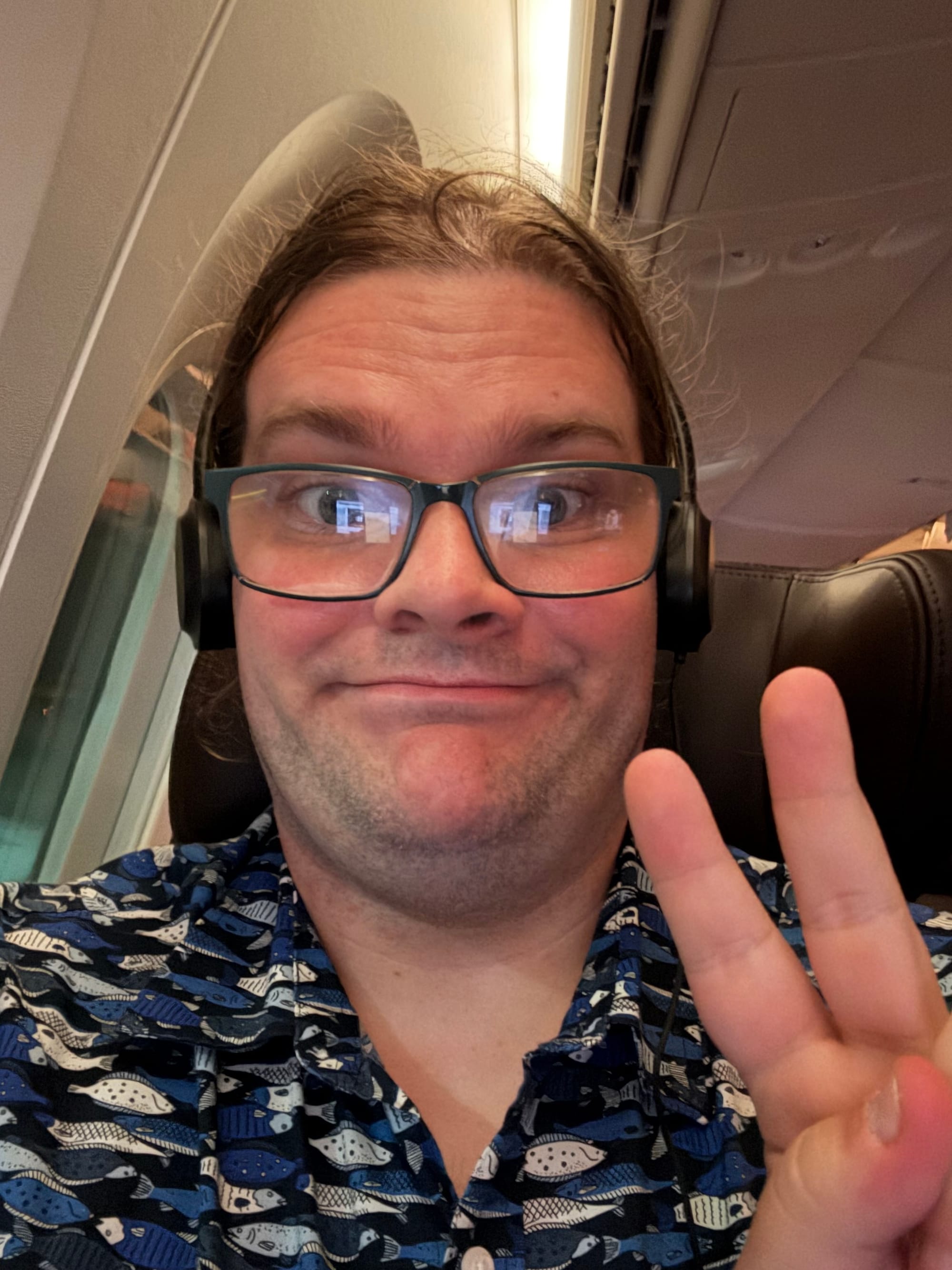 Traveller wearing glasses and headphones gives a peace sign while seated by the window on an Etihad Airways flight, shortly after departure from Manchester Airport.