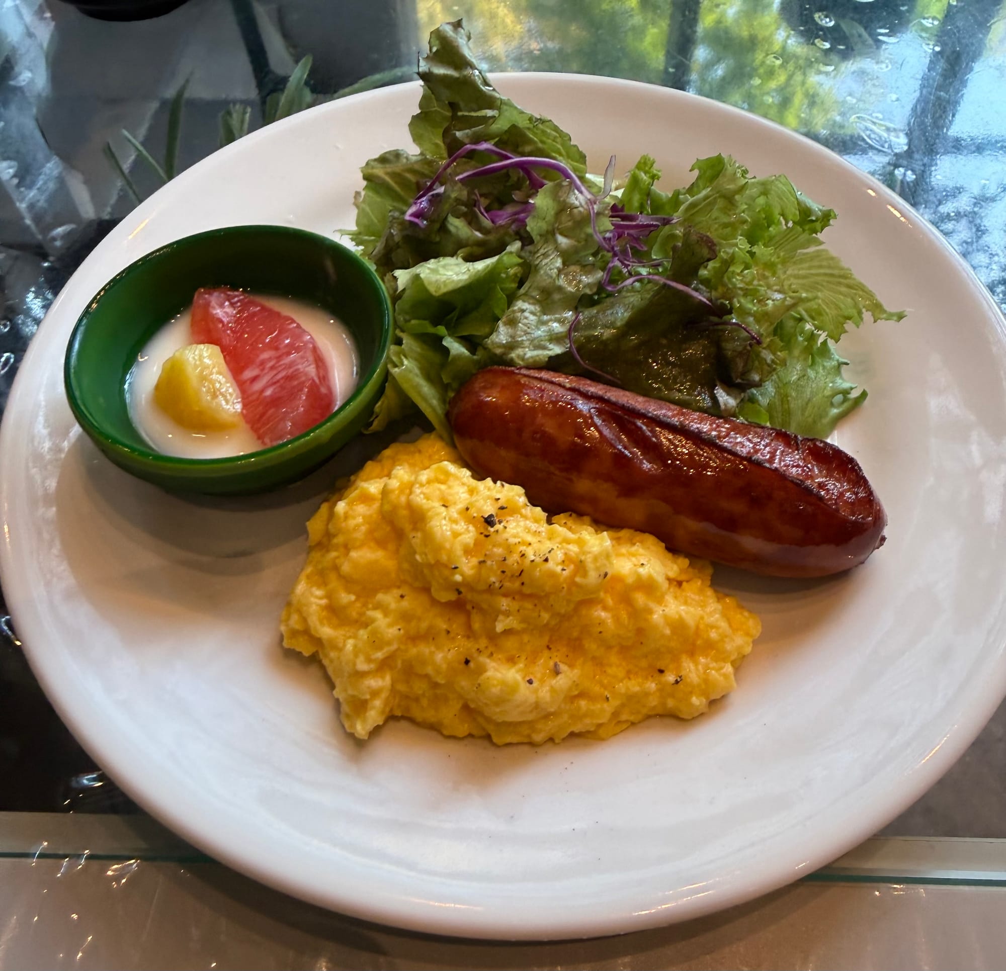 Breakfast plate with scrambled eggs, a grilled sausage, mixed salad and a small fruit yoghurt, served on a white plate in a hotel restaurant.