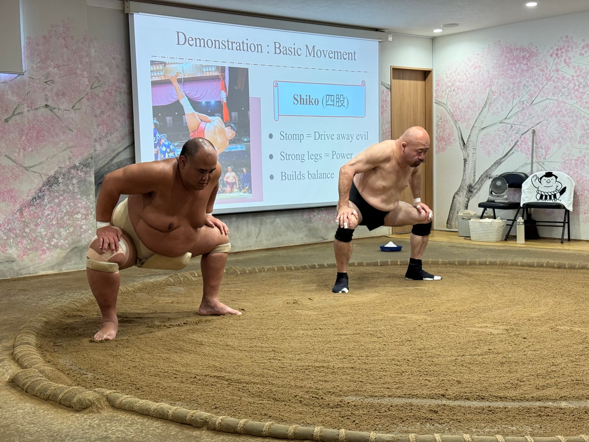 Two sumo wrestlers demonstrate the traditional shiko exercise inside a dojo in Osaka, standing in the ring before a presentation explaining its purpose and technique.