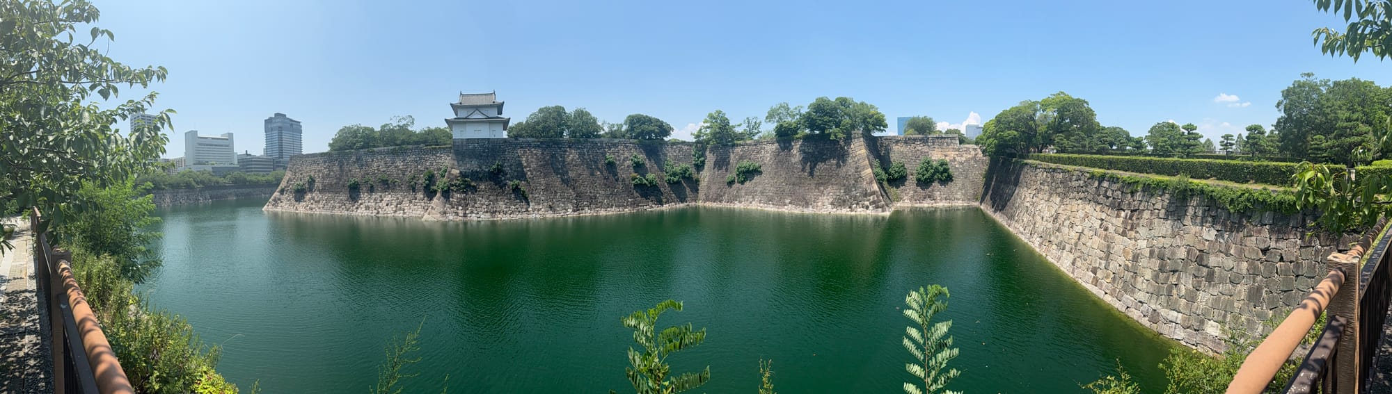 Panoramic view of Osaka Castle's moat, with emerald-green water surrounded by towering stone walls, a watchtower and trees beneath a clear blue summer sky.