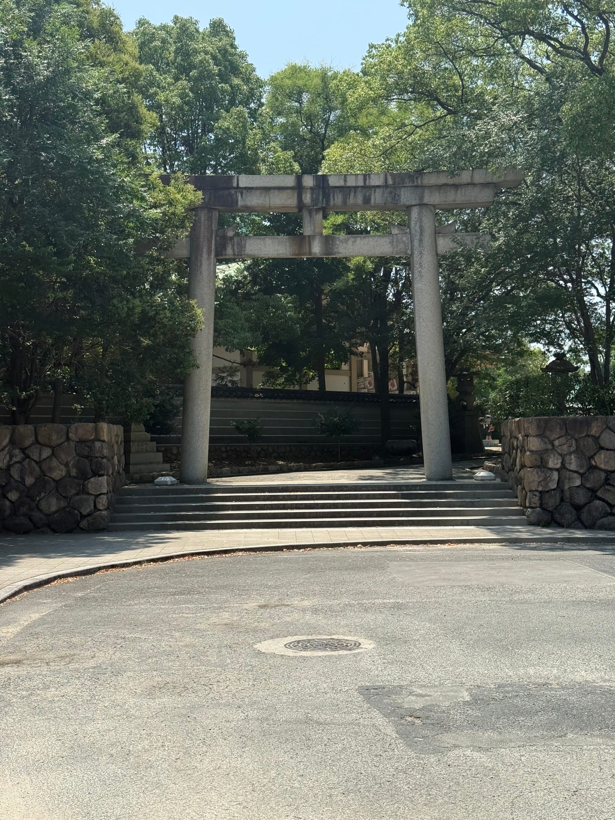 Stone torii gate within Osaka Castle Park, framed by mature trees and stone walls, marking the entrance to a Shinto shrine beneath a bright summer sky.