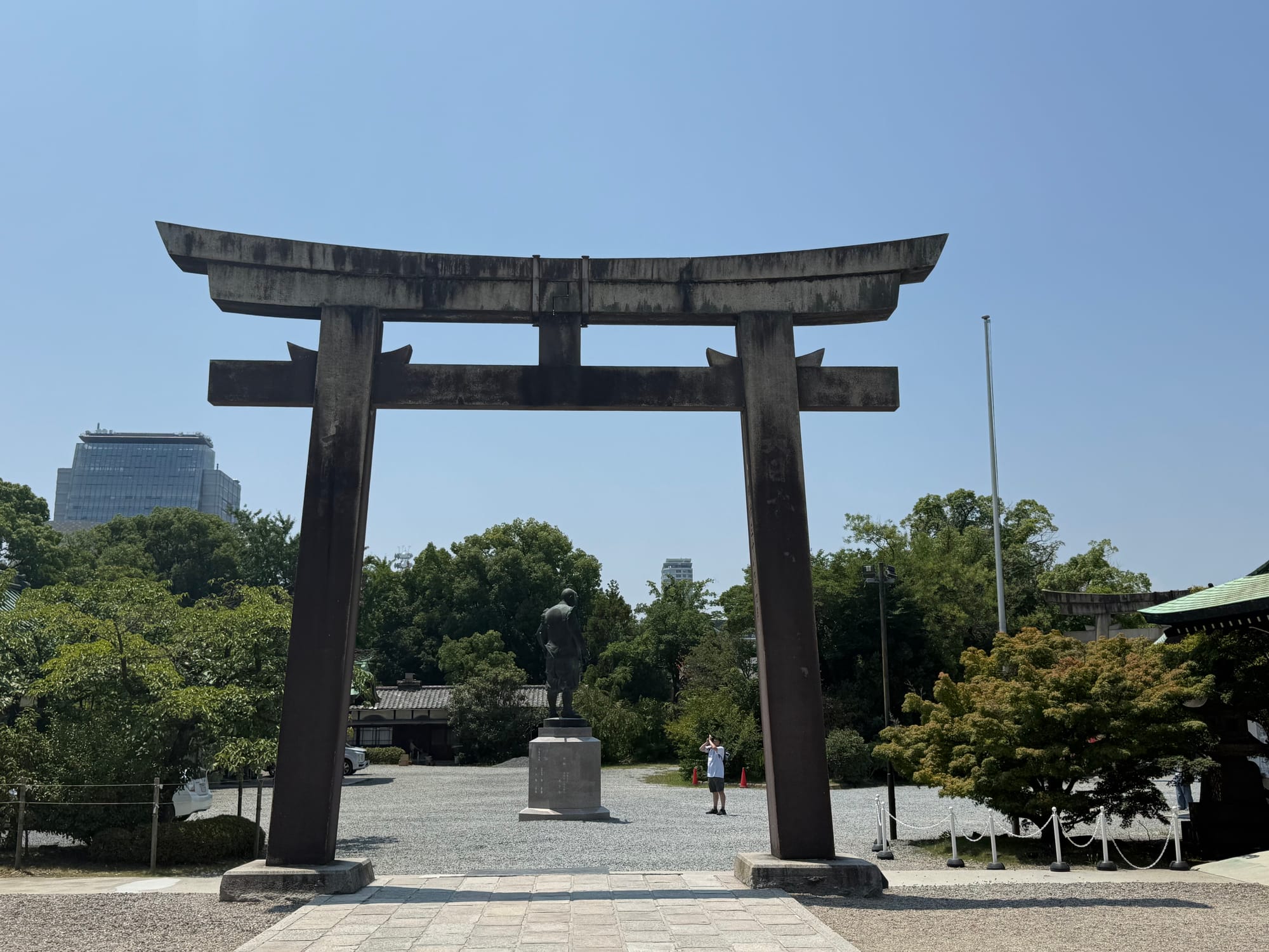 Large stone torii gate in Osaka Castle Park, standing before a statue and shrine grounds, framed by trees and viewed beneath a clear blue summer sky.