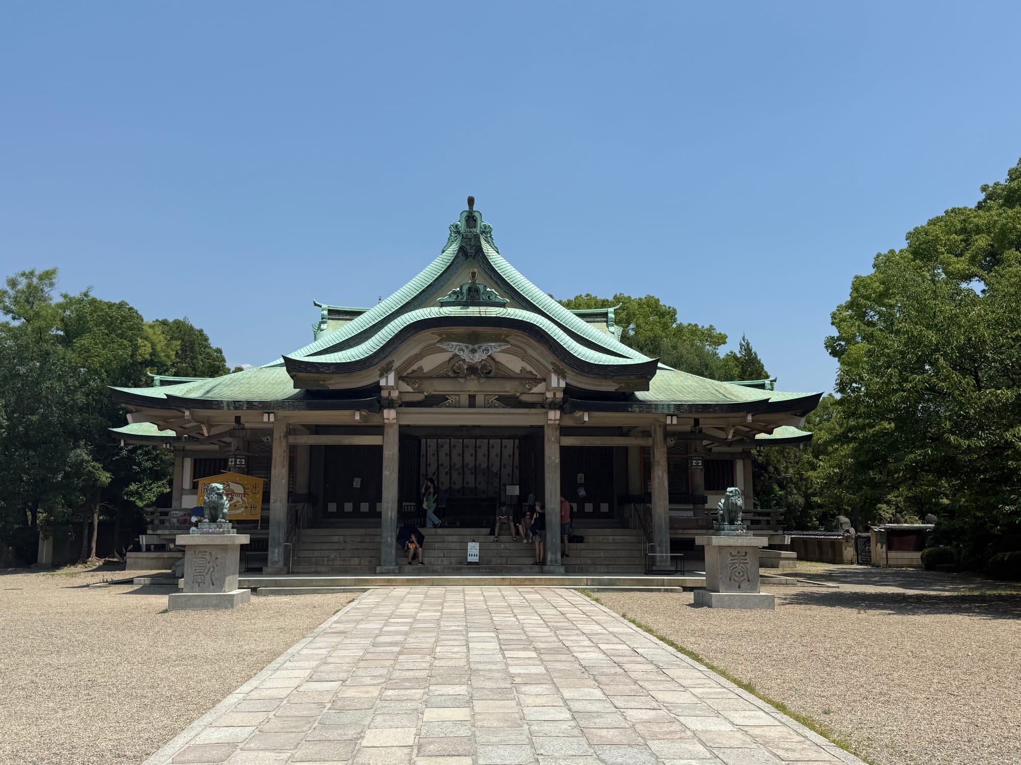 Hokoku Shrine in Osaka Castle Park, featuring a traditional Shinto shrine building with a curved copper-green roof, stone pathway and guardian statues beneath a clear blue sky.