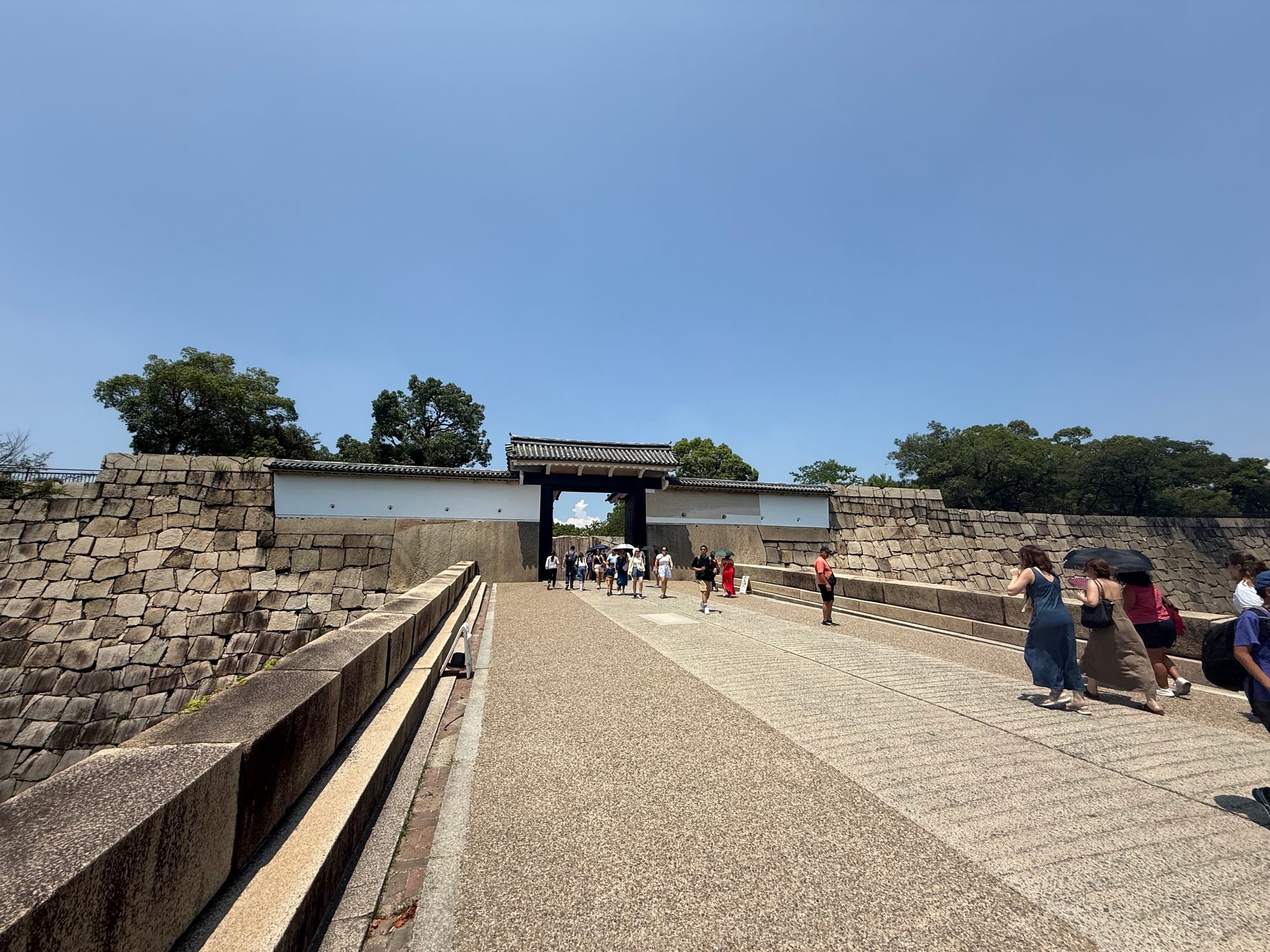 Sakuramon Gate at Osaka Castle, viewed across a broad stone bridge and surrounded by towering defensive walls, with visitors entering beneath a clear summer sky.