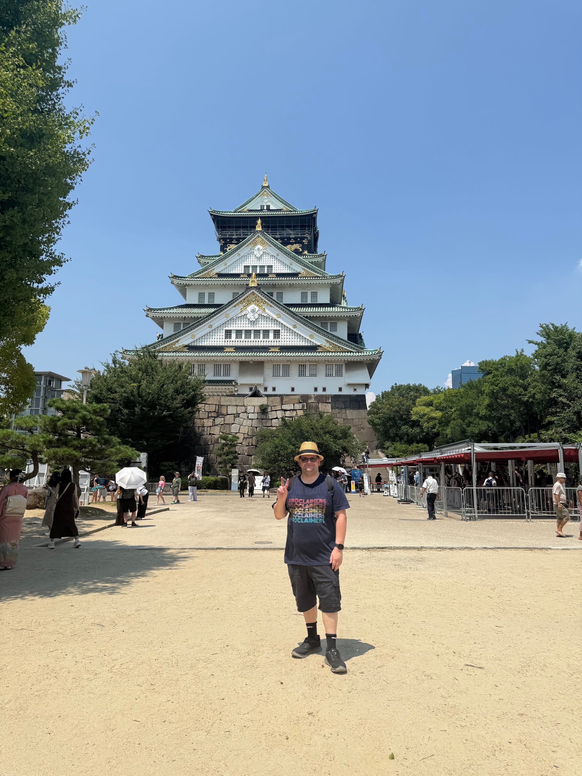Visitor standing in front of Osaka Castle, making a peace sign, with the historic castle keep rising above stone walls and trees beneath a clear summer sky.