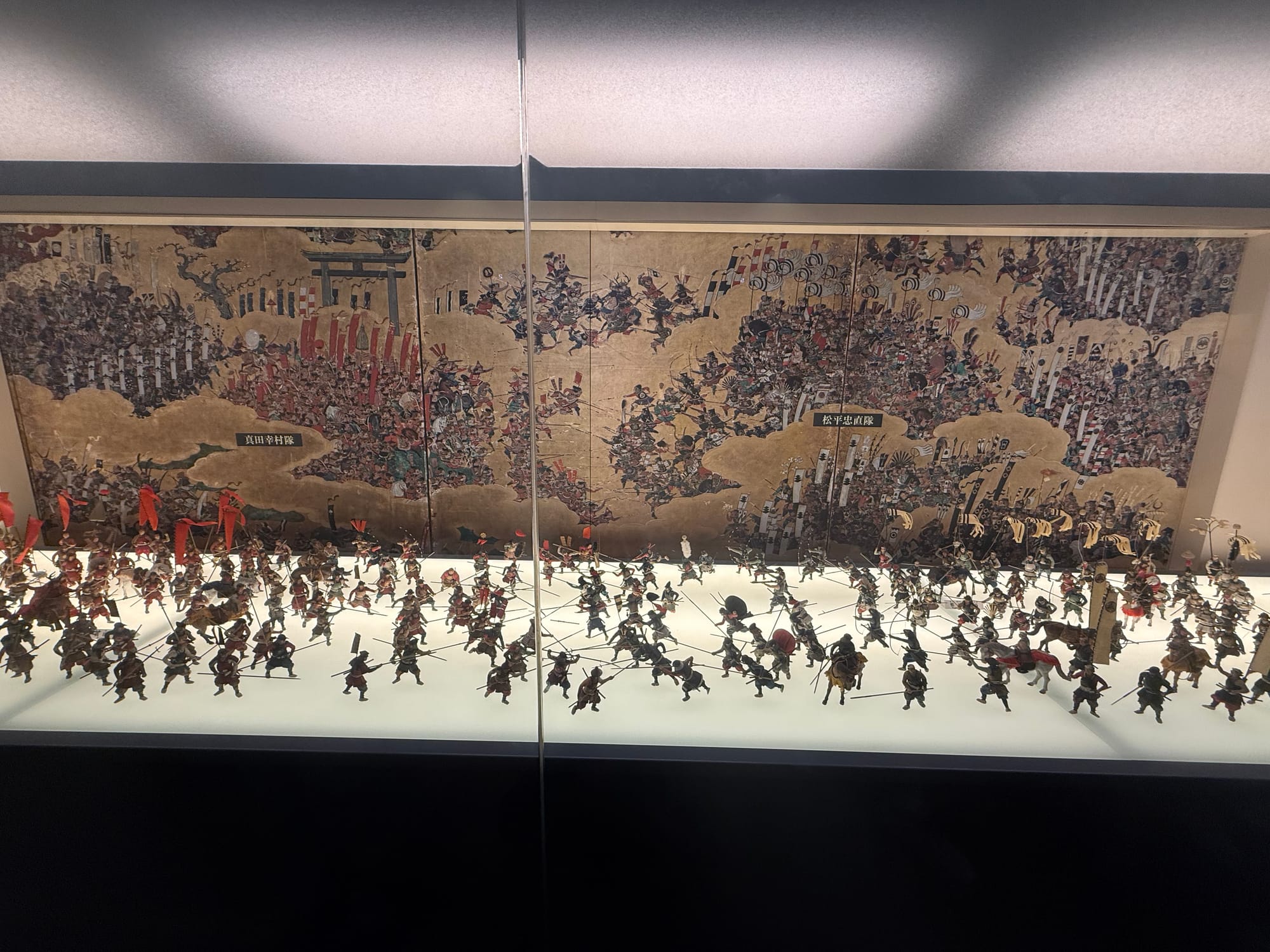 Diorama of samurai warriors and cavalry displayed in Osaka Castle Museum, set before a detailed battlefield screen depicting fighting during the Siege of Osaka.