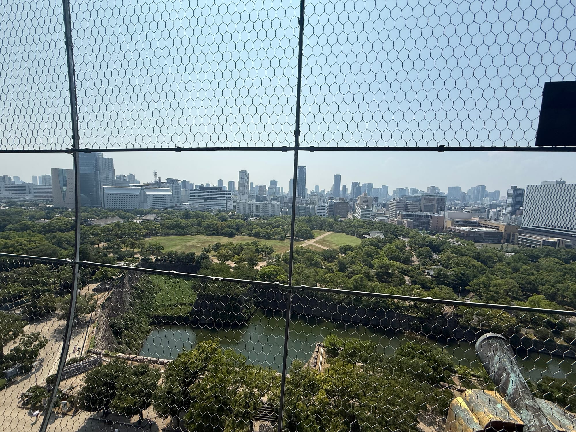 View west from Osaka Castle's observation deck across the castle moat and parkland towards central Osaka, with modern skyscrapers stretching across the skyline.