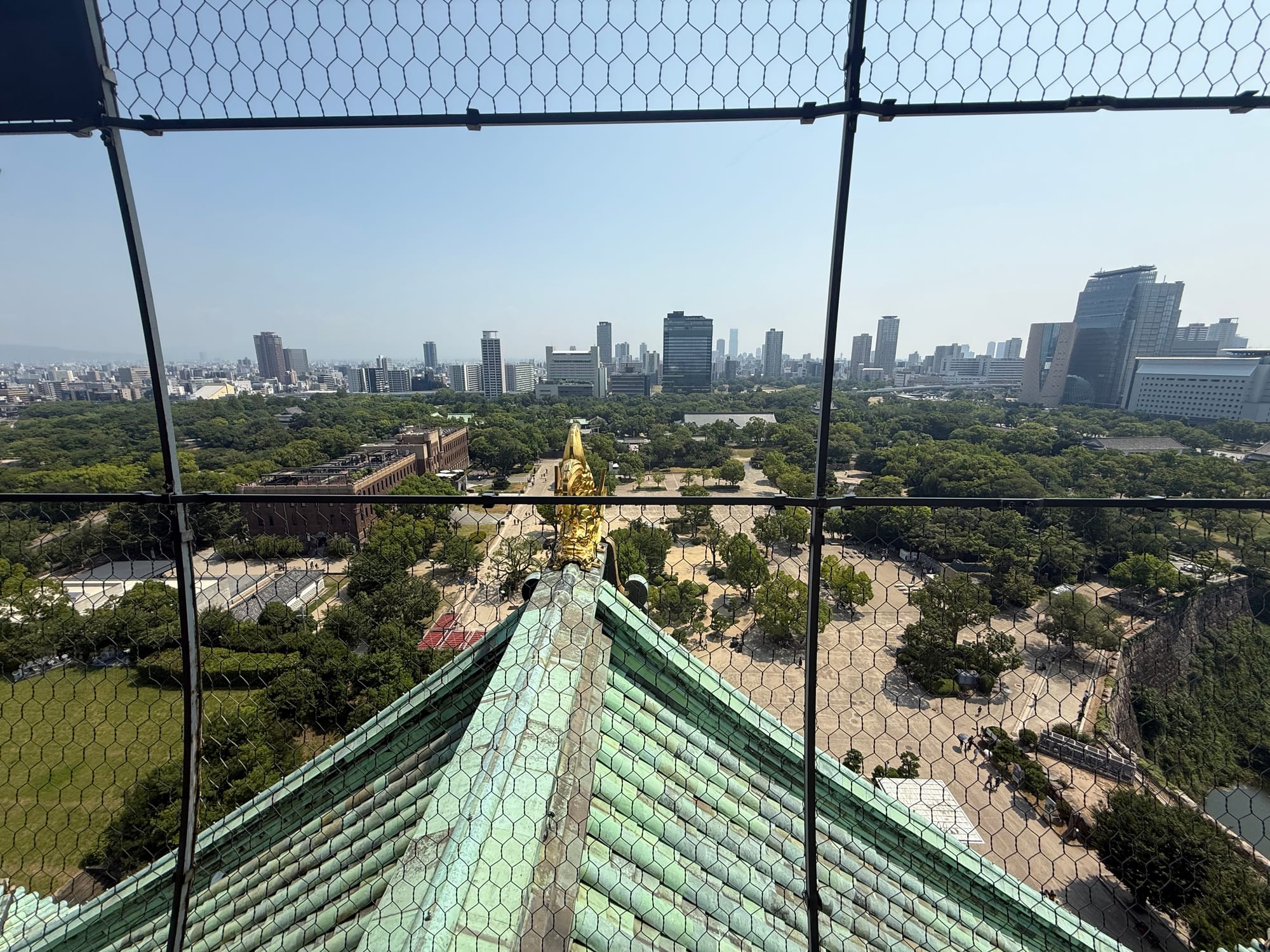 View south from Osaka Castle's observation deck, looking over the castle roof with a golden shachihoko ornament towards Osaka Castle Park and the modern city skyline.