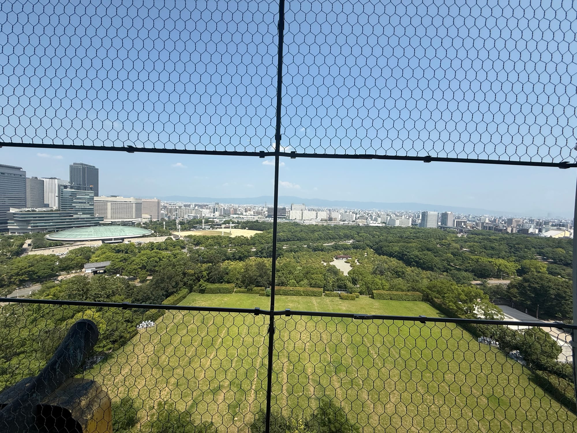 View east from Osaka Castle's observation deck across castle parkland and moats towards central Osaka, with office towers, city streets and distant mountains on the horizon.