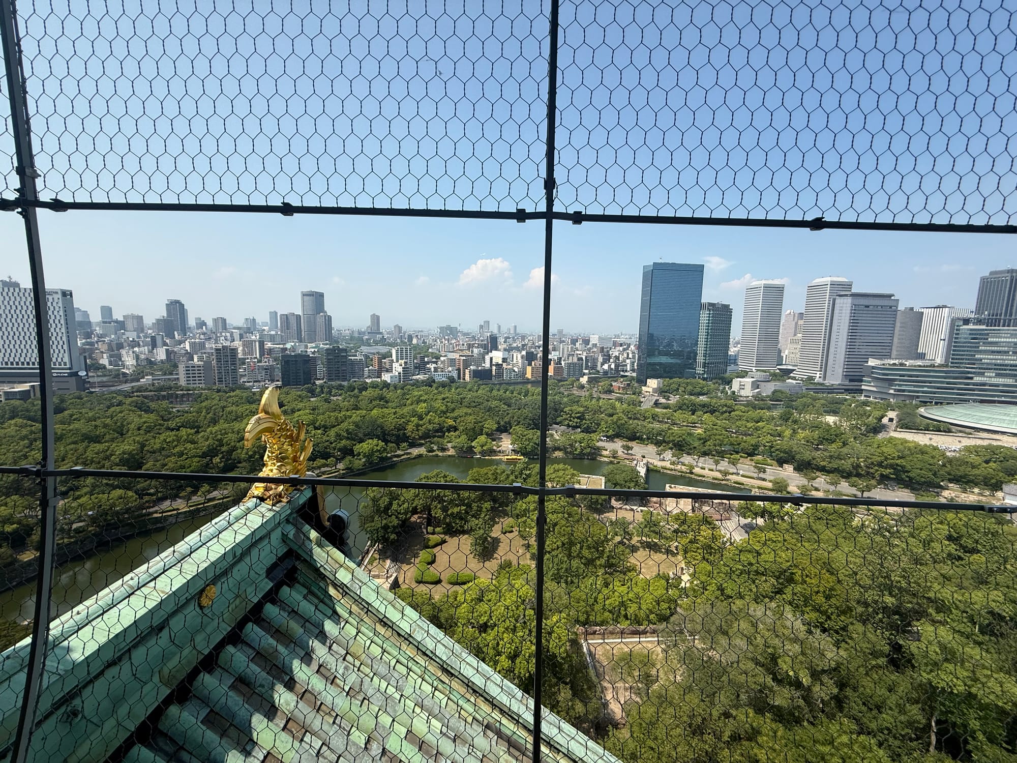 View north from Osaka Castle's observation deck across the castle moat and parkland, with a golden shachihoko roof ornament and Osaka's modern skyline beyond.