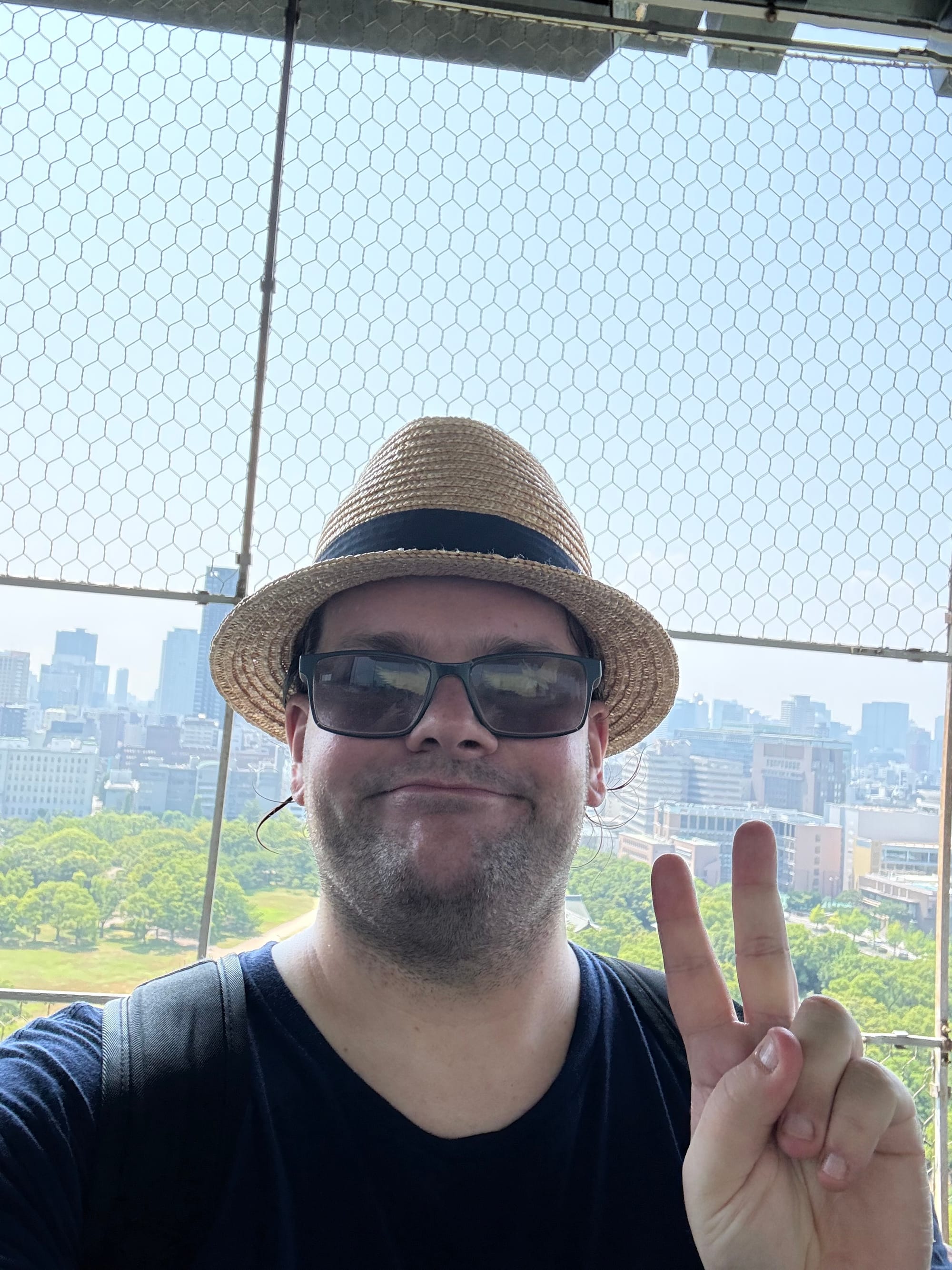Visitor wearing a straw hat and sunglasses poses for a selfie on the observation deck of Osaka Castle, making a peace sign with the city skyline visible in the background.