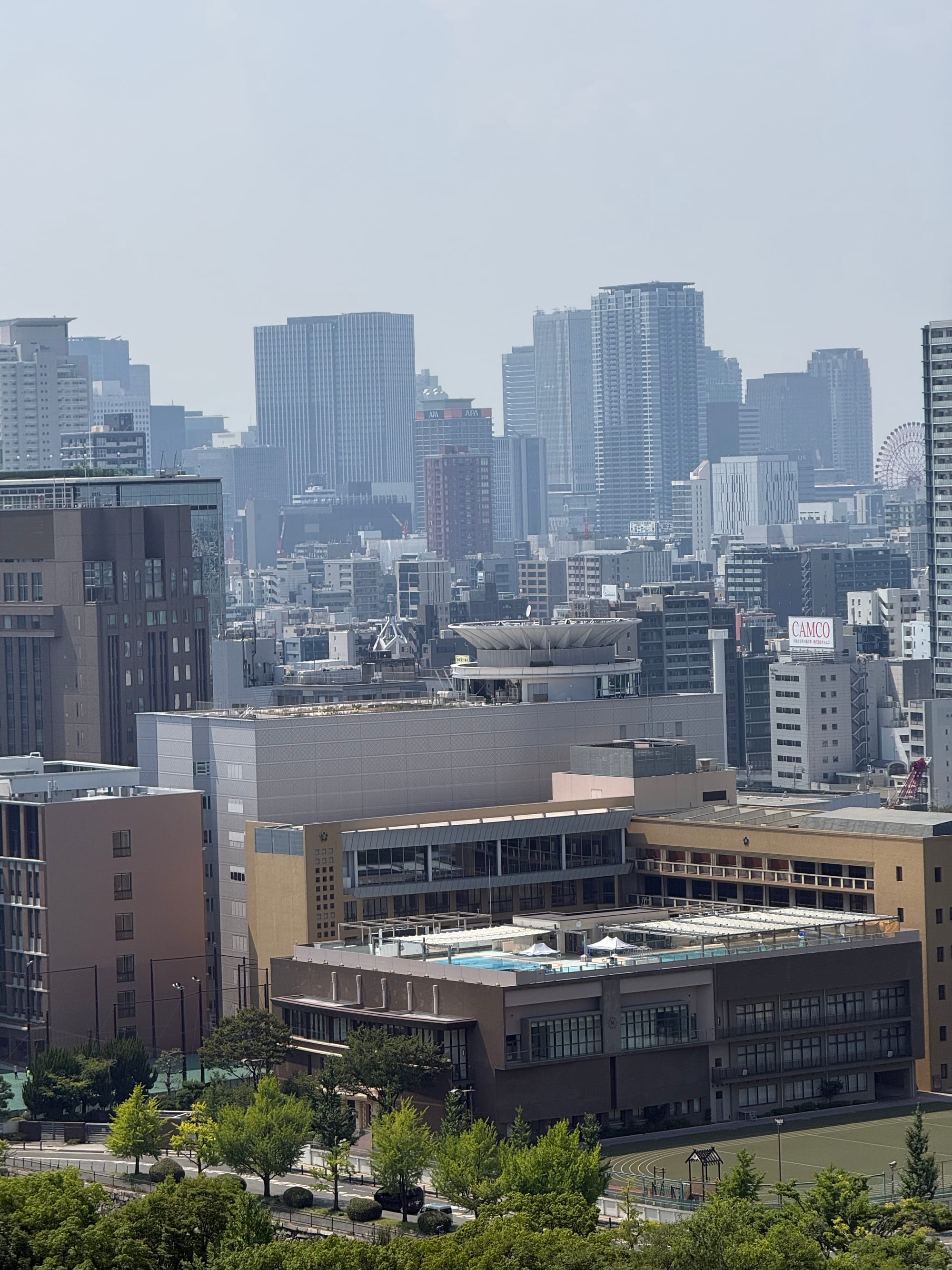 View across central Osaka from Osaka Castle, showing modern office towers, apartment blocks and city streets stretching towards the horizon on a hazy summer day.