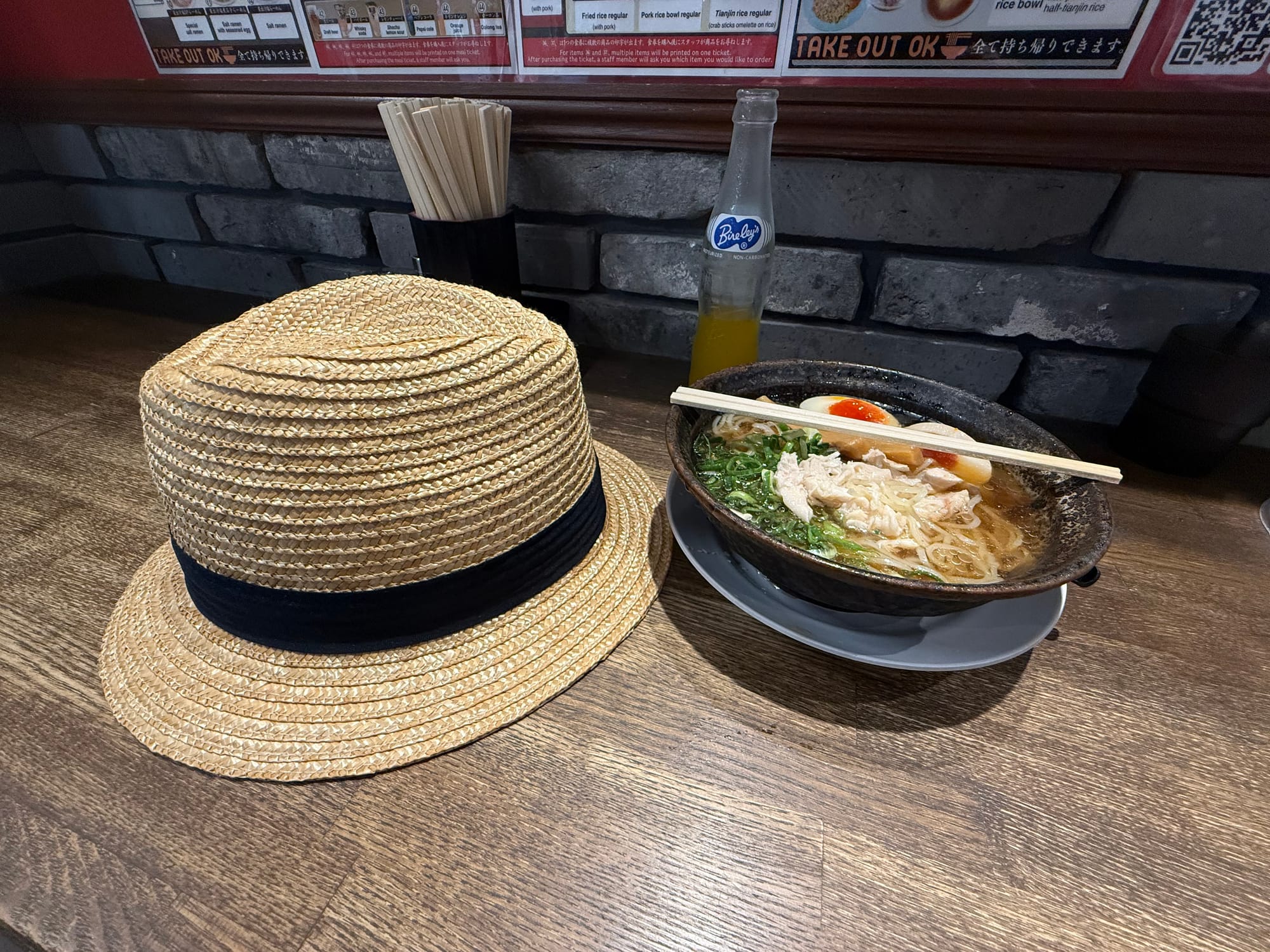 Straw hat resting beside a bowl of ramen topped with sliced pork, spring onions and egg, served at a counter in a Japanese restaurant.