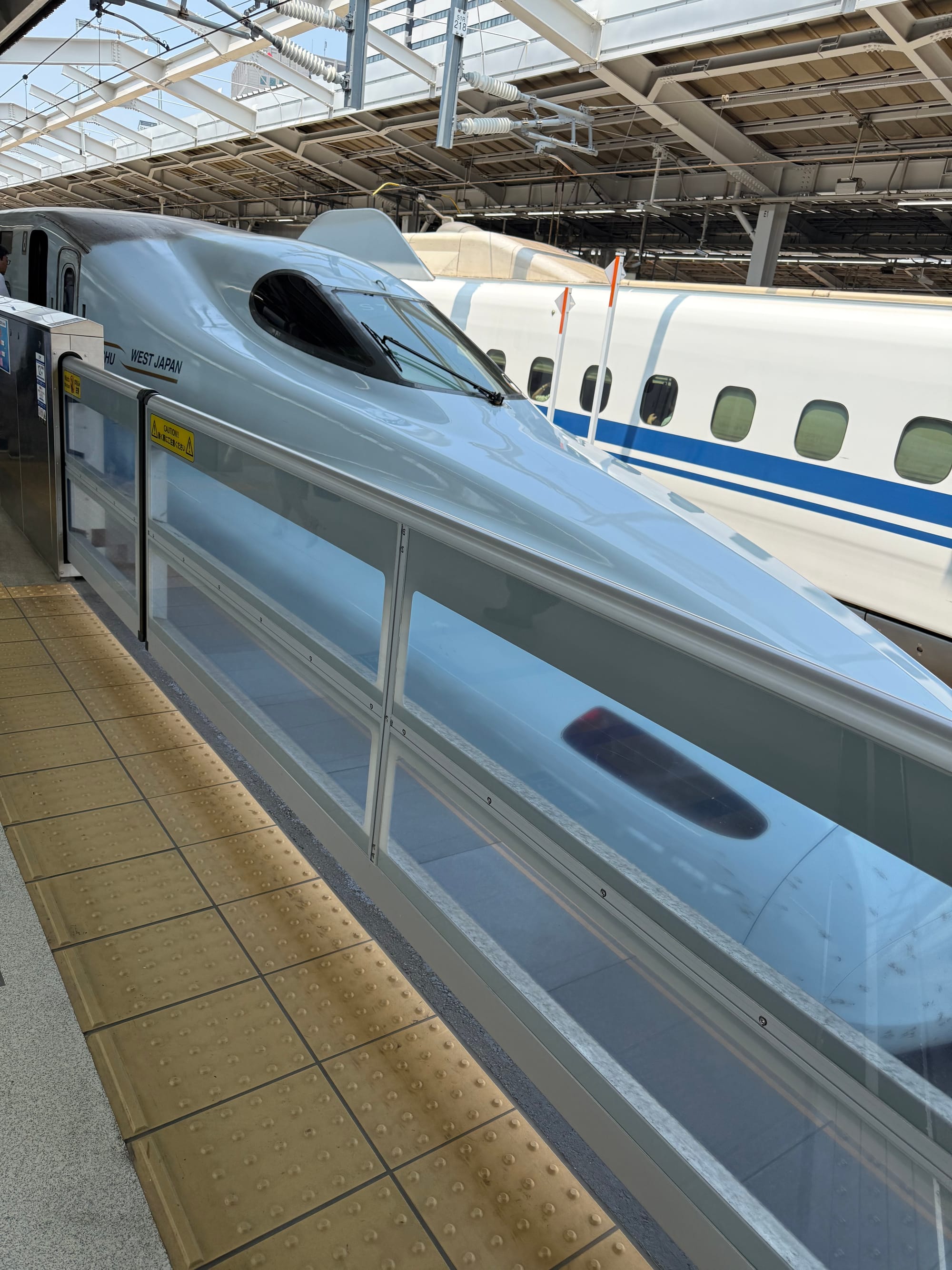 JR West N700 Series Shinkansen at a station platform, showing its streamlined nose and aerodynamic design beneath the station canopy and overhead infrastructure.