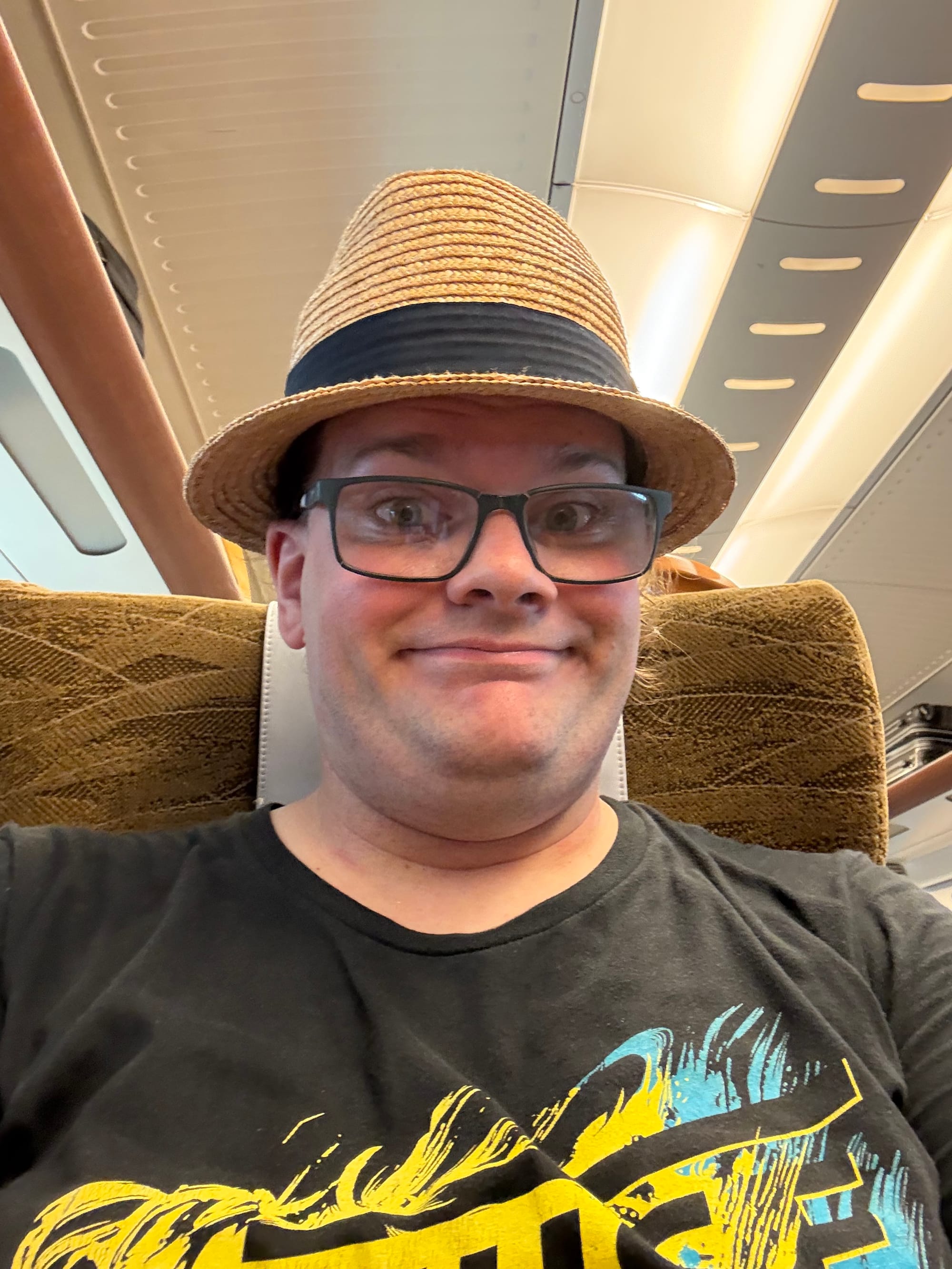 Traveller wearing a straw hat and glasses takes a selfie while seated on a Shinkansen, with patterned seats and the train's interior visible in the background.