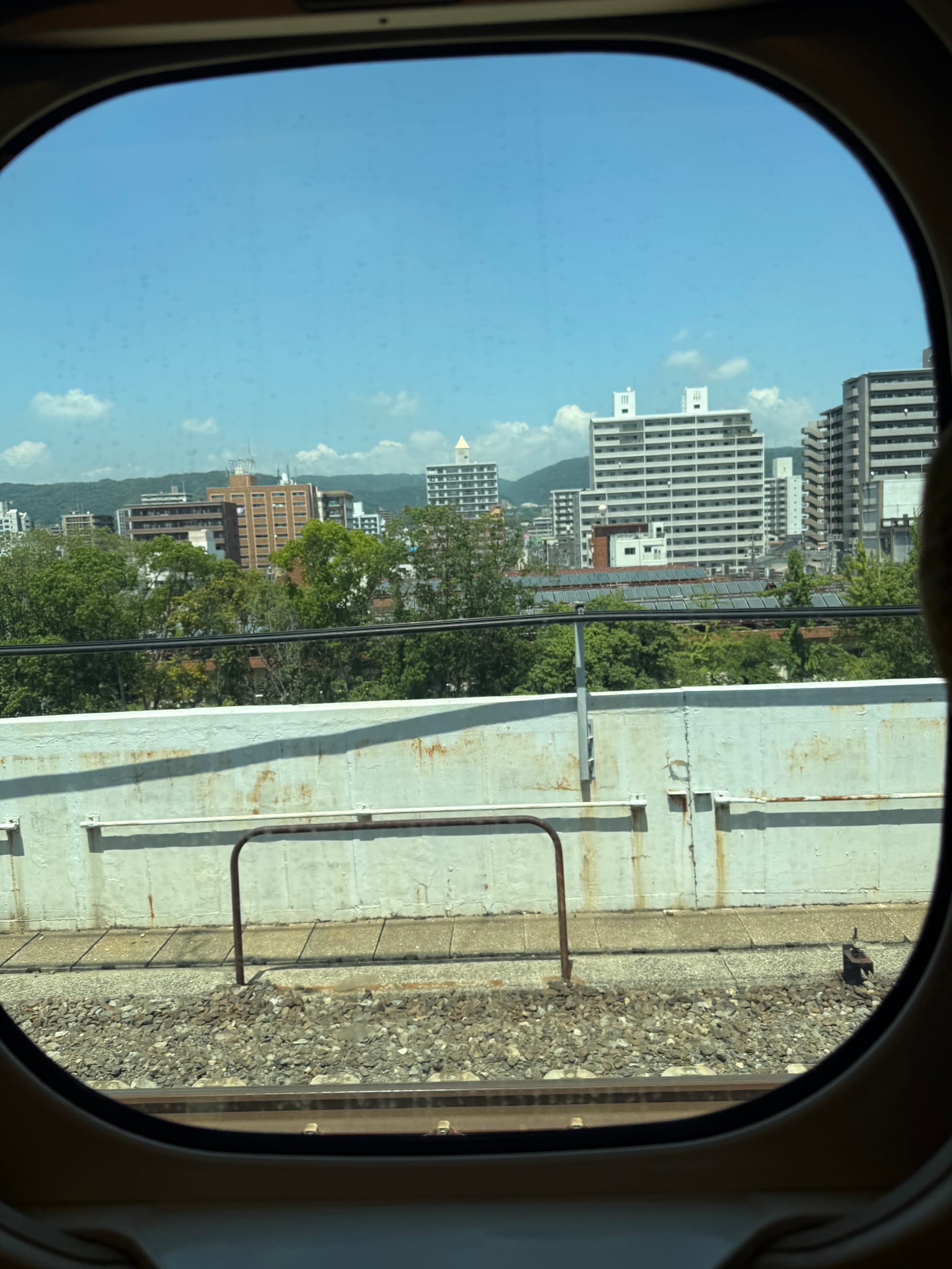 View from a Shinkansen window showing apartment blocks, trees and distant hills beneath a clear blue sky, framed by the train's distinctive rounded window.