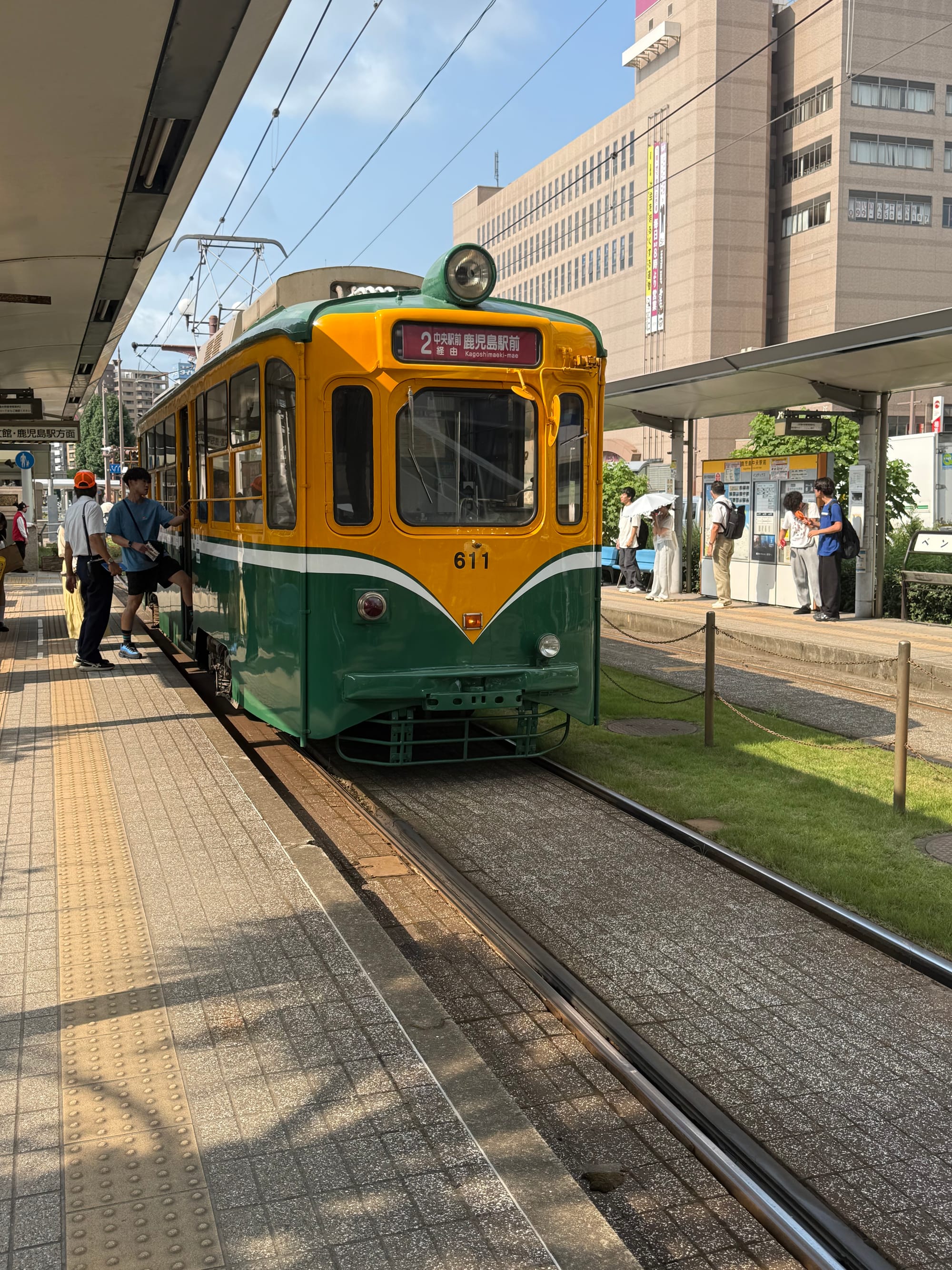 Green and yellow Kagoshima City Tram 611 waiting at a stop outside Kagoshima-Chūō Station, with passengers boarding beneath a bright summer sky.