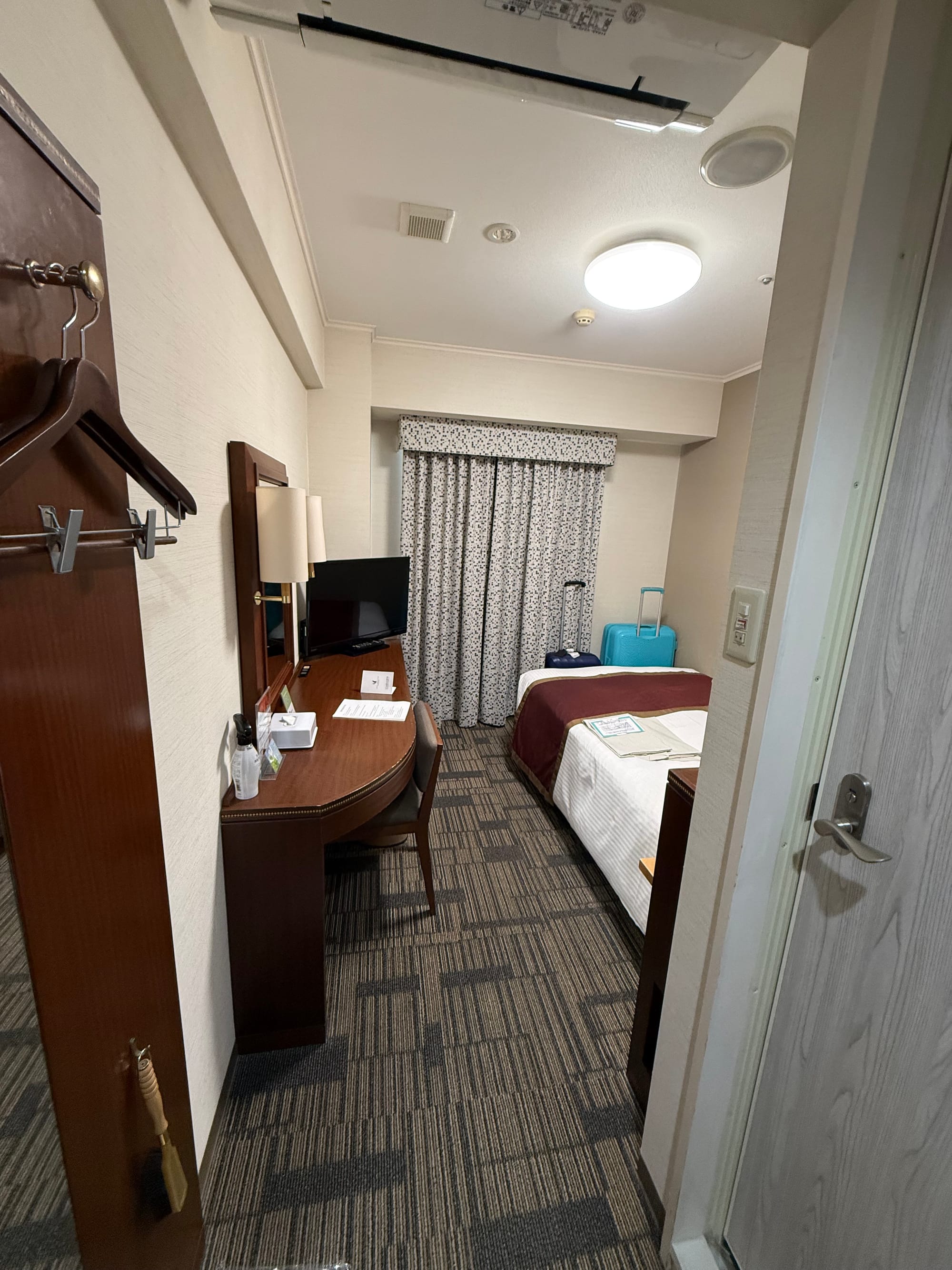 Compact hotel room in Kagoshima featuring a single bed, desk, television, luggage and patterned curtains, viewed from the entrance doorway.