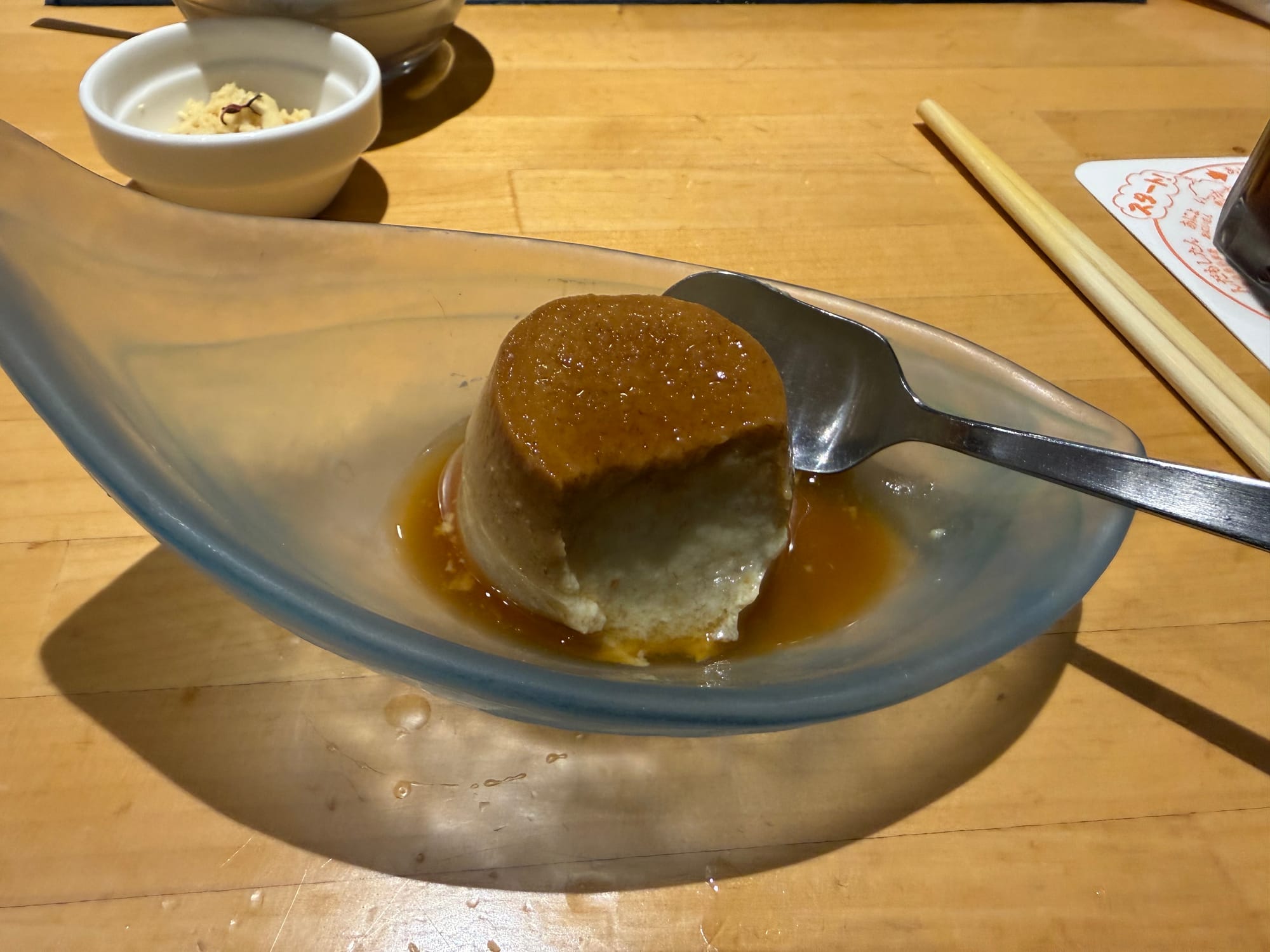 Banana-flavoured pudding dessert served in a glass dish with caramel sauce, partially eaten and shown with a spoon after a meal in a Kagoshima restaurant.