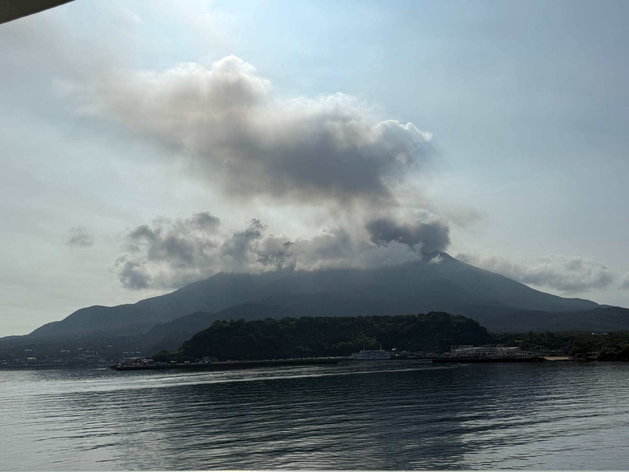 Sakurajima volcano rises above calm waters near Kagoshima, with volcanic clouds and steam drifting from the summit beneath a hazy afternoon sky.