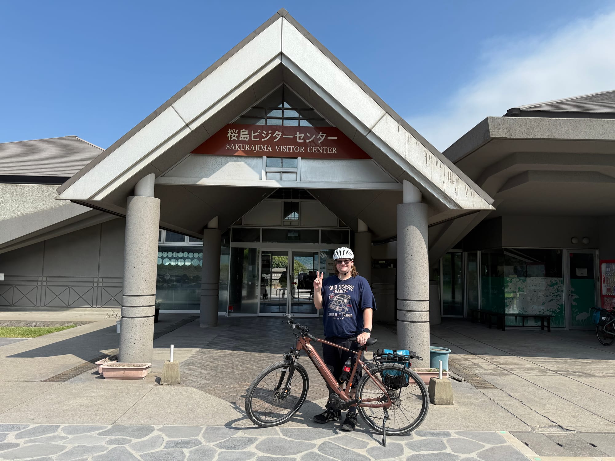 Visitor stands with a bicycle outside the Sakurajima Visitor Centre, wearing a cycling helmet and making a peace sign beneath the entrance sign on a sunny day.