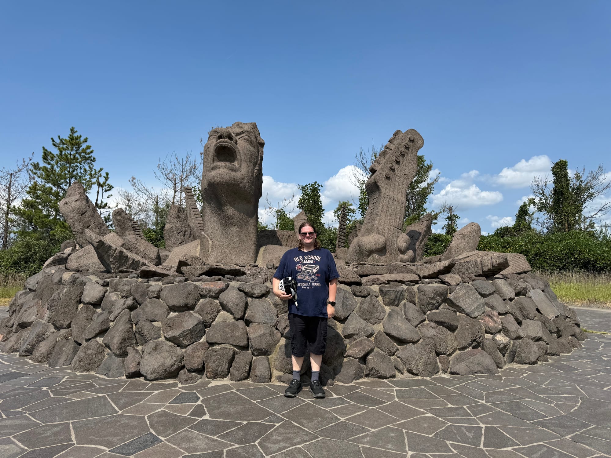 Visitor stands beside the "Portrait of Scream" lava sculpture on Sakurajima, featuring expressive carved faces and abstract volcanic rock forms beneath a clear blue sky.
