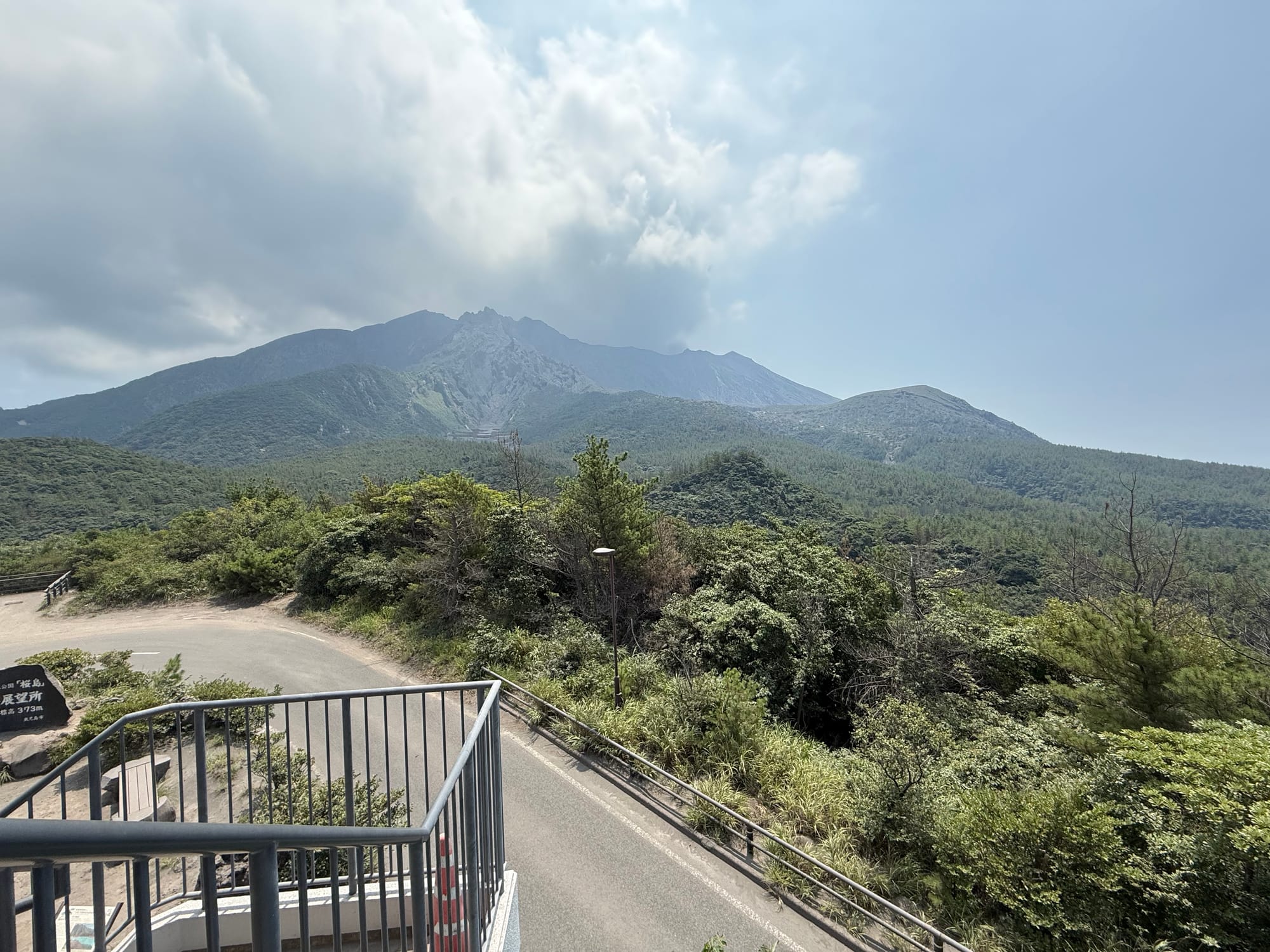 View from Yunohira Observatory on Sakurajima, looking across forested slopes towards the volcano's rugged upper crater area beneath cloud and drifting volcanic haze.