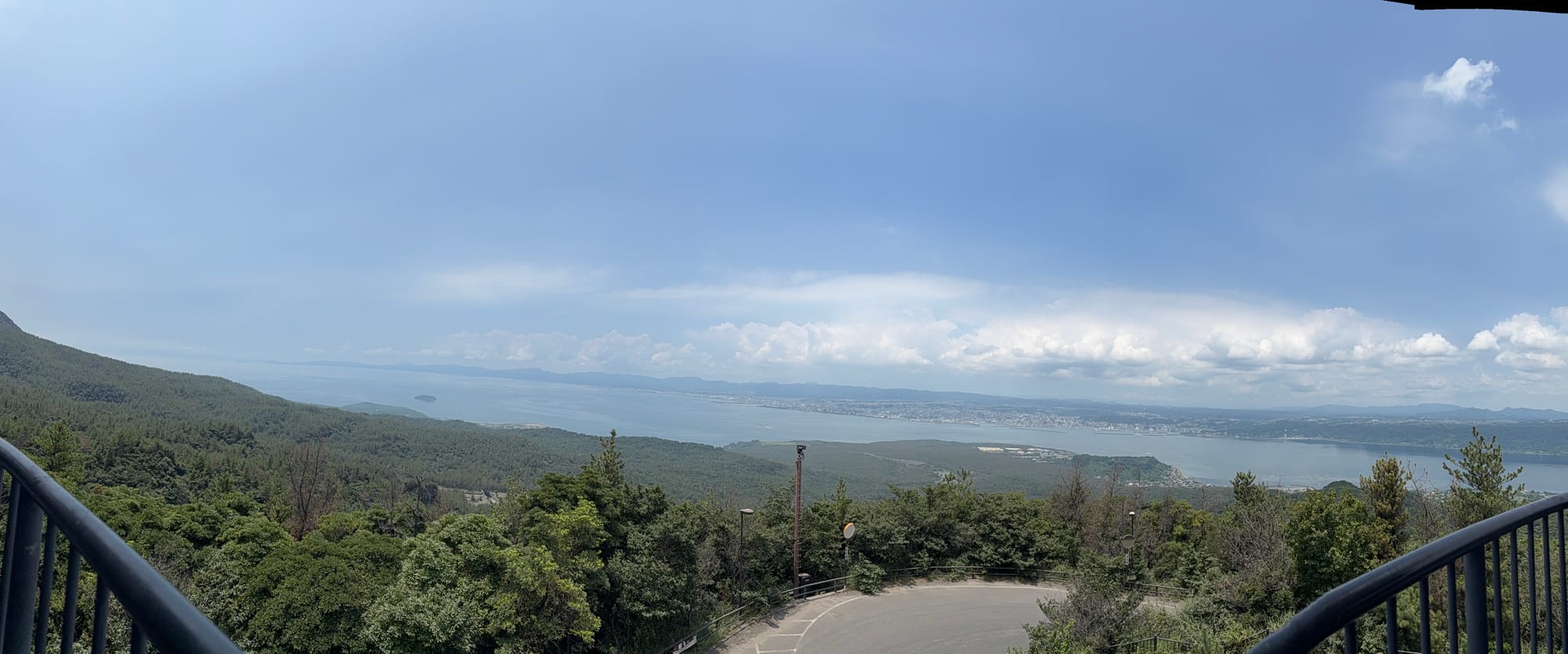 View from Yunohira Observatory on Sakurajima across forested slopes and Kagoshima Bay, with the city of Kagoshima visible beneath dramatic summer clouds.