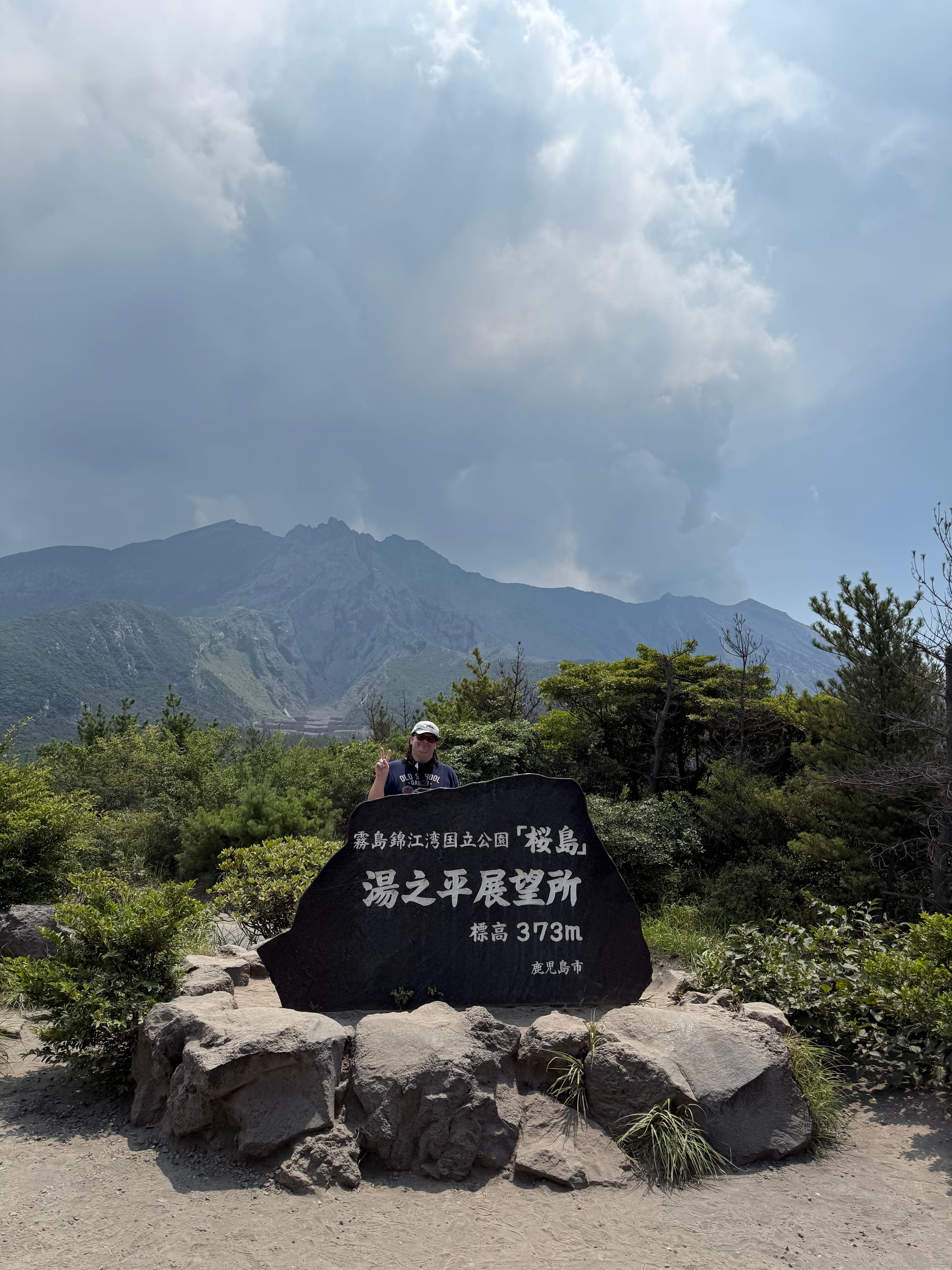 Visitor stands behind a sign at Yunohira Observatory on Sakurajima, with the volcanic slopes and crater area rising behind beneath cloud and volcanic haze.