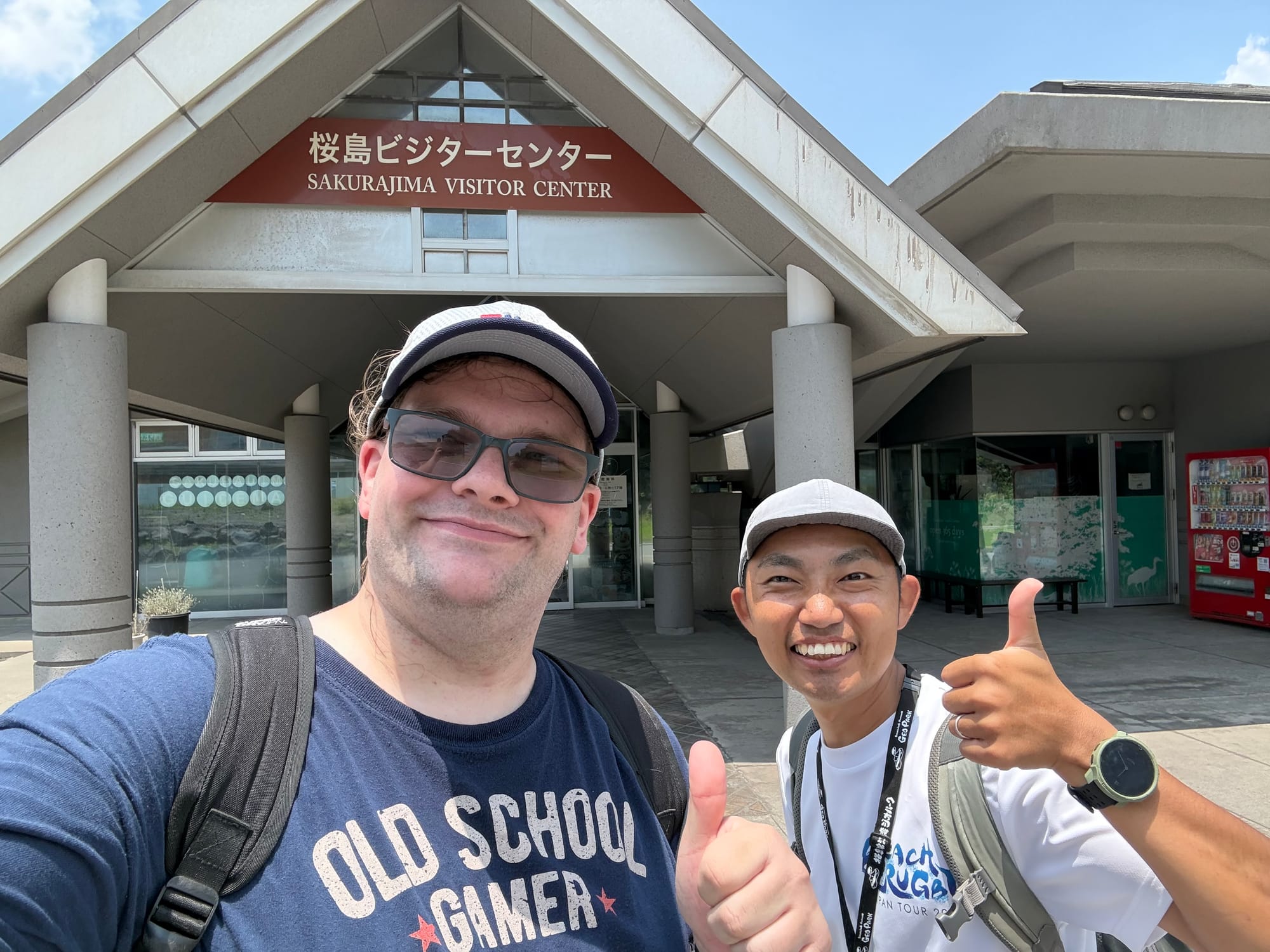 Two people give thumbs up outside the Sakurajima Visitor Centre, standing beneath the entrance sign on a bright summer day during a cycling tour of the island.