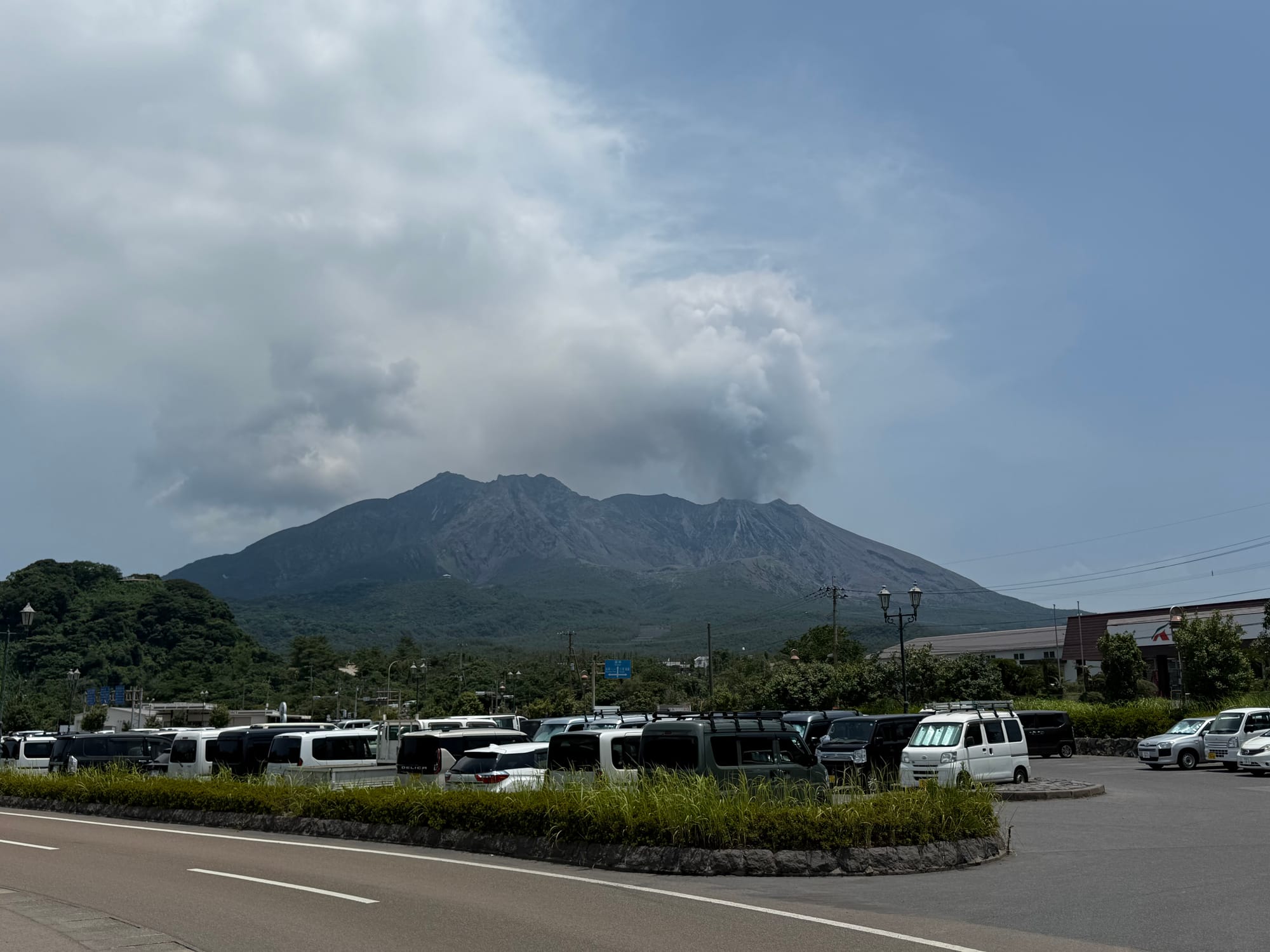 Sakurajima volcano viewed from the island, with a plume of volcanic ash rising from the summit above parked vehicles and greenery beneath a bright summer sky.