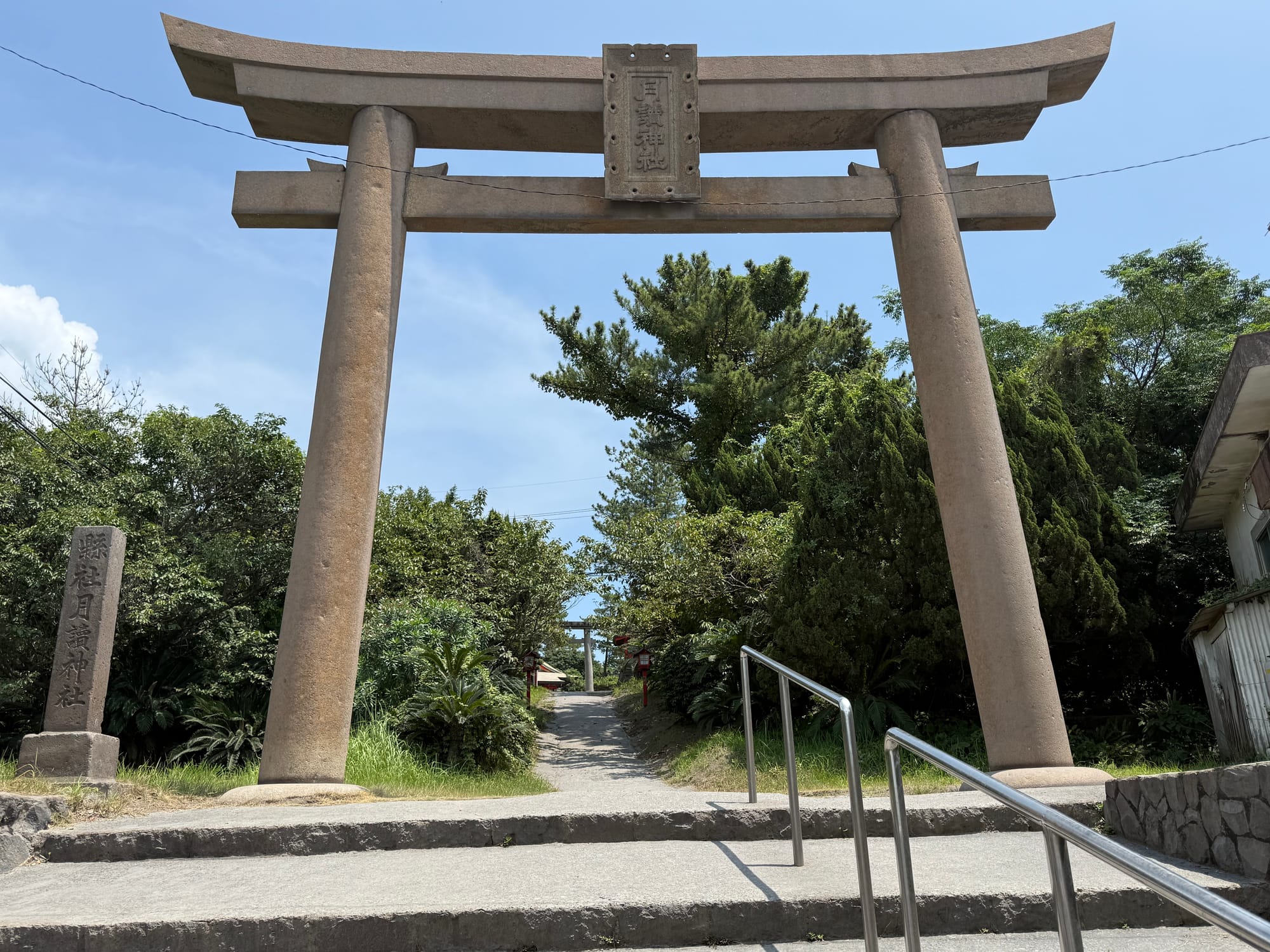 Large stone torii gate at Tsukiyomi Shrine on Sakurajima, standing above steps leading to a shaded pathway lined with trees and traditional shrine structures.