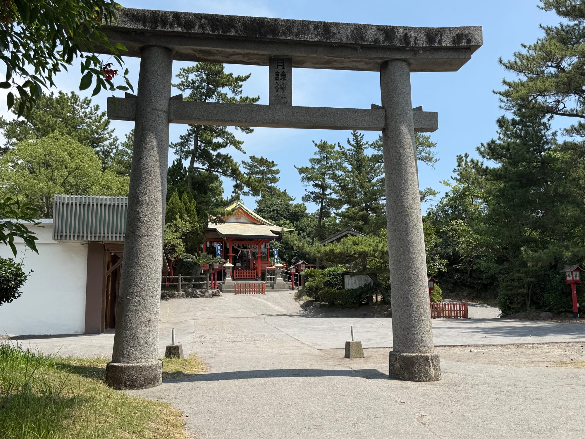 Stone torii gate at Tsukiyomi Shrine on Sakurajima frames the shrine buildings beyond, surrounded by pine trees, lanterns and peaceful shrine grounds.