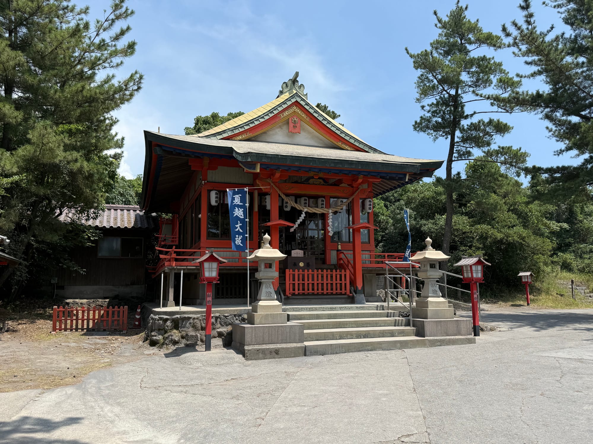 Main hall of Tsukiyomi Shrine on Sakurajima, featuring red-painted wooden architecture, stone lanterns, banners and sacred Shinto decorations among pine trees.