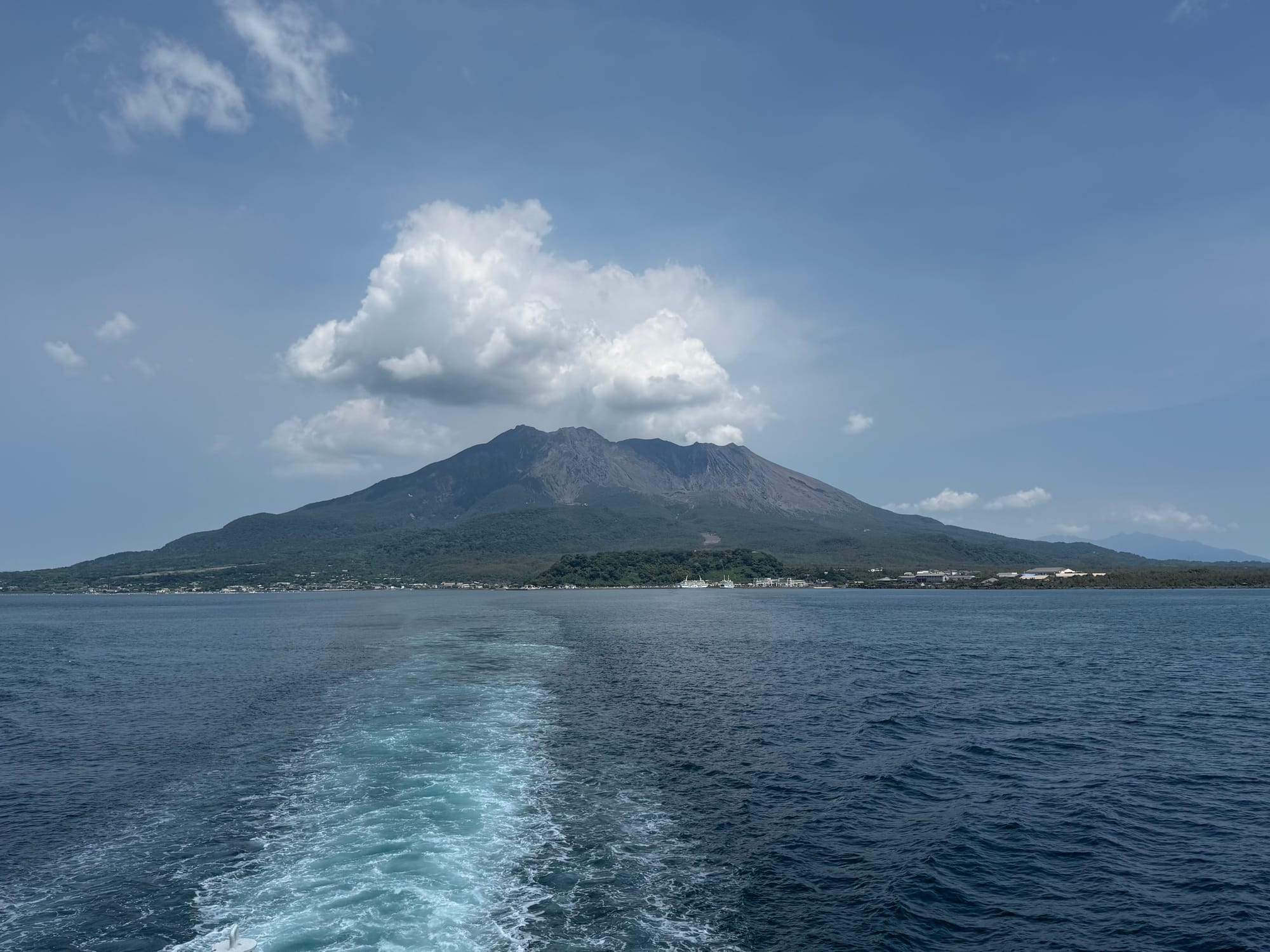 View from a ferry departing Sakurajima, with the volcanic island rising above Kagoshima Bay and the vessel's wake stretching across the water beneath a summer sky.